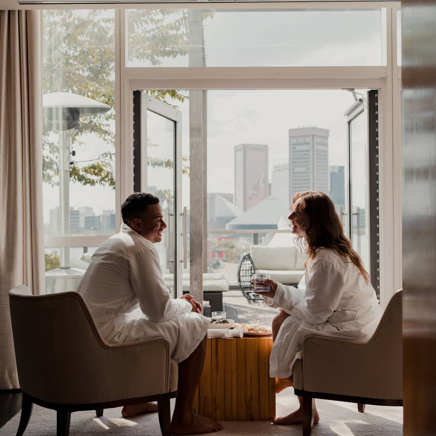 Two people in white robes sit in armchairs by an open balcony door with a cityscape view in the background, sharing a relaxed, casual conversation.