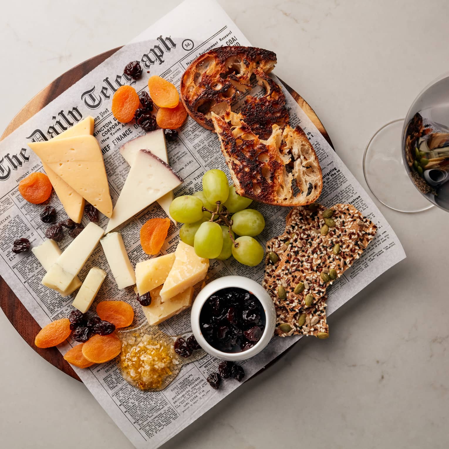 Cheese, grapes, crackers and bread on newspaper on platter next to glass of red wine