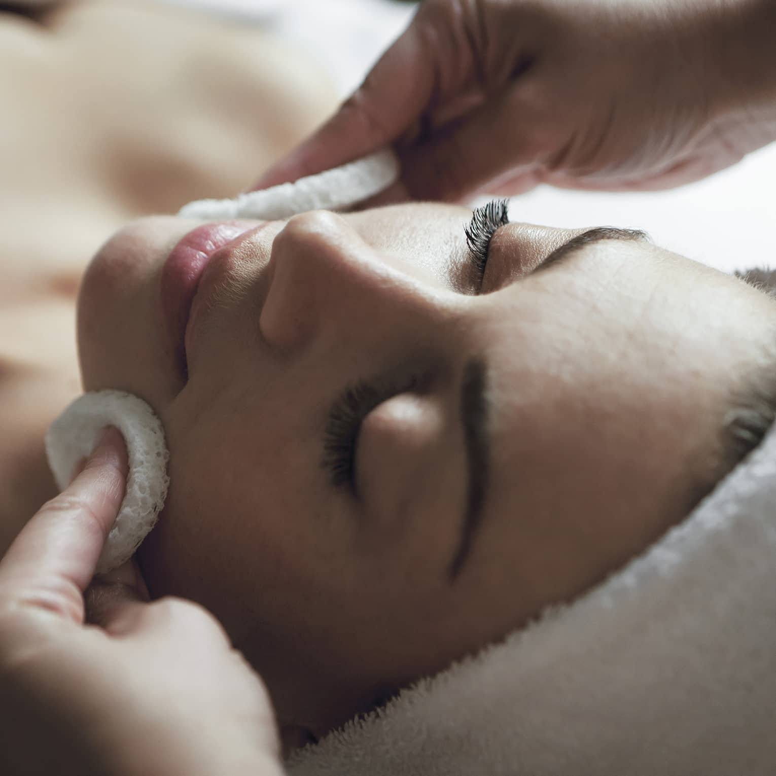 A detail of hands rubbing a sponge on a woman's face as she lies on a massage table with her eyes closed and hair wrapped up in a white towel
