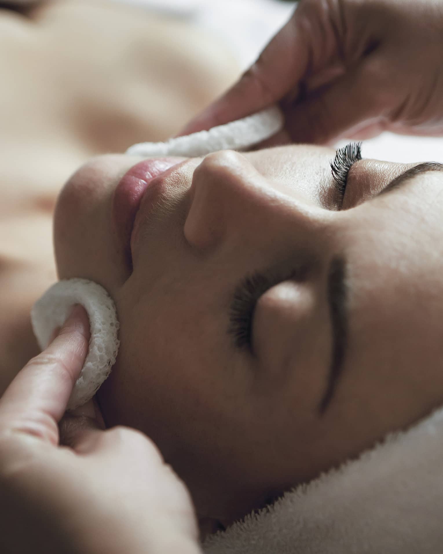 A detail of hands rubbing a sponge on a woman's face as she lies on a massage table with her eyes closed and hair wrapped up in a white towel