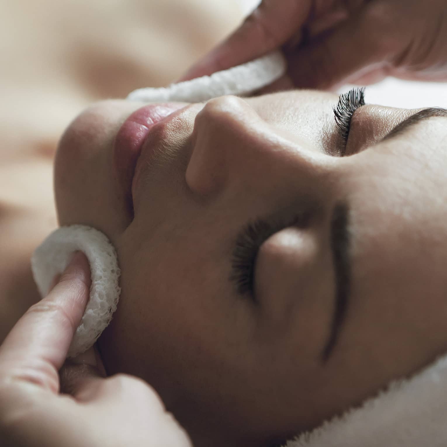 A detail of hands rubbing a sponge on a woman's face as she lies on a massage table with her eyes closed and hair wrapped up in a white towel