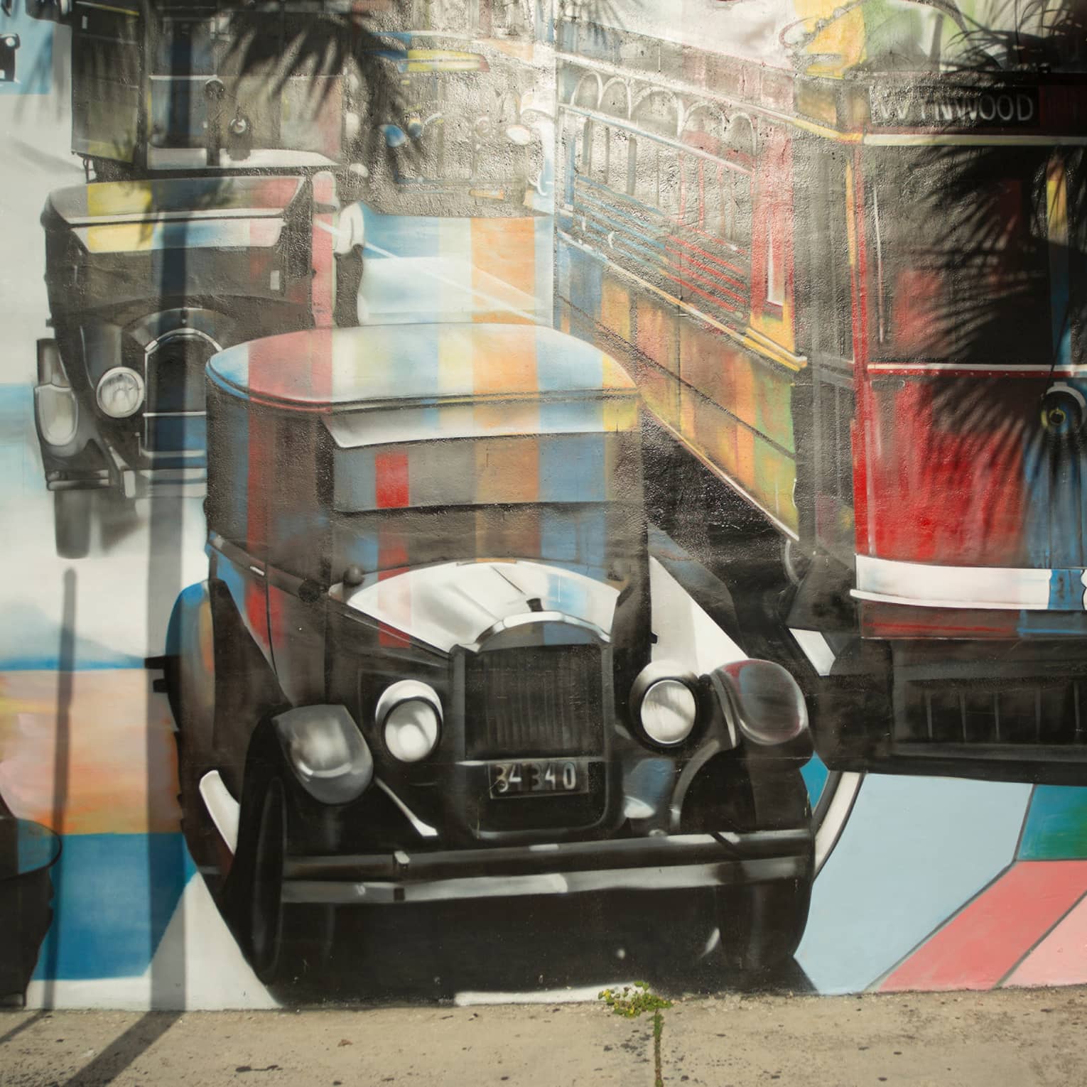 Palm trees cast shadows on a mural of vintage cars and a bus painted in a kaleiscopic pattern of colours and grey shading.