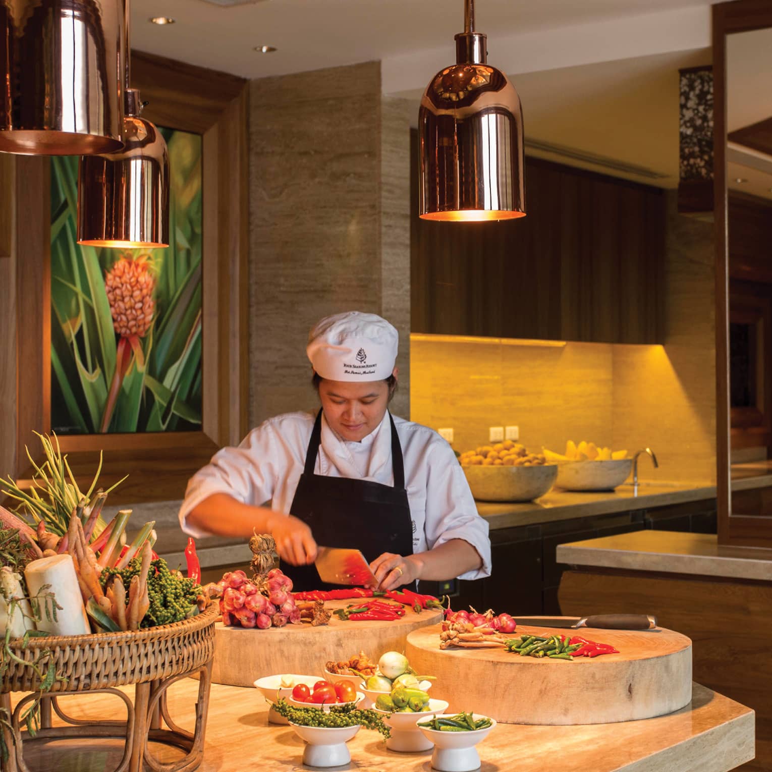 A chef chops vegetables on a large butcher block amid a vibrant array of fresh produce in a luxurious, warm-hued kitchen.