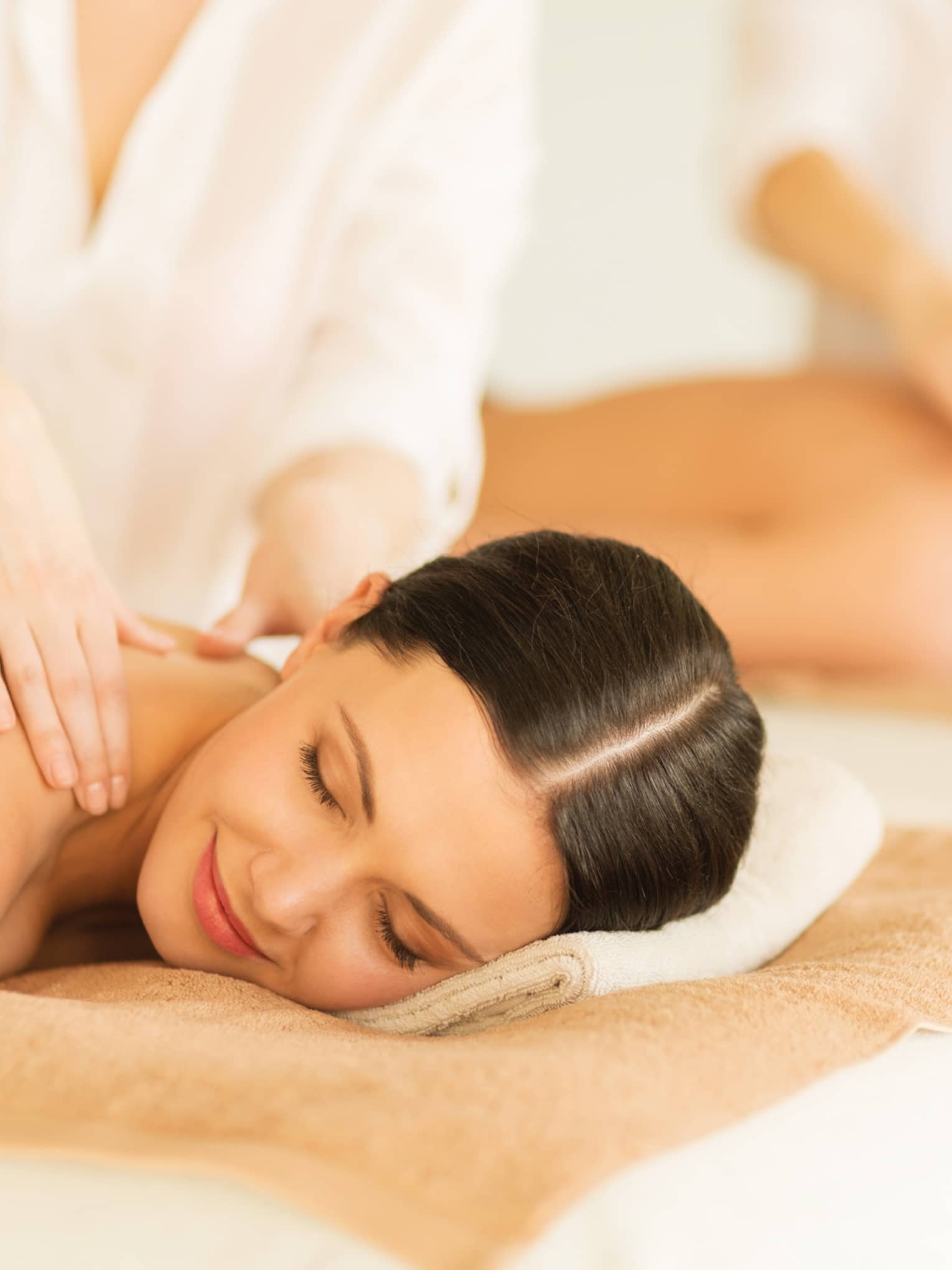 Woman and man lie side-by-side on massage tables as spa staff massage their bare shoulders