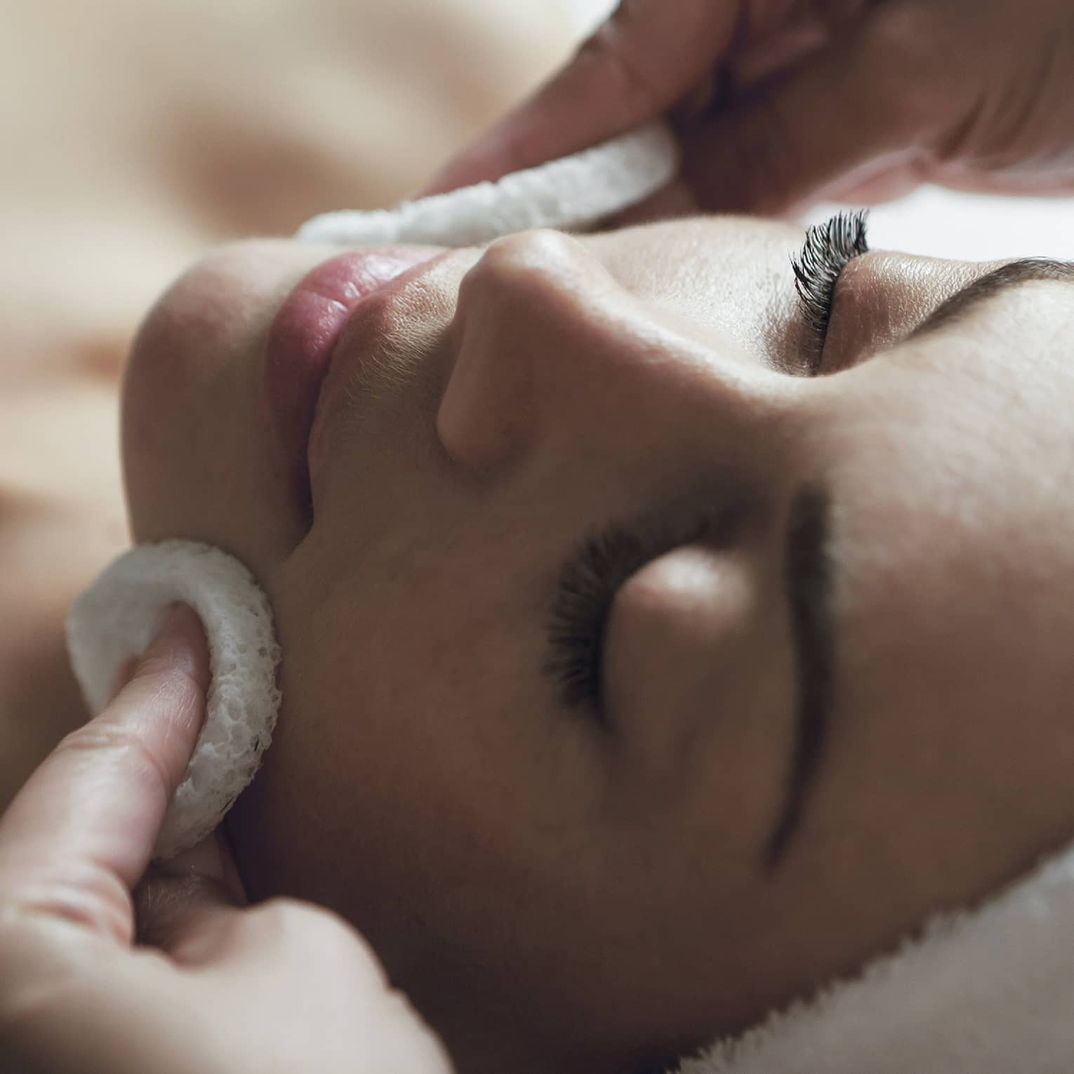 A detail of hands rubbing a sponge on a woman's face as she lies on a massage table with her eyes closed and hair wrapped up in a white towel