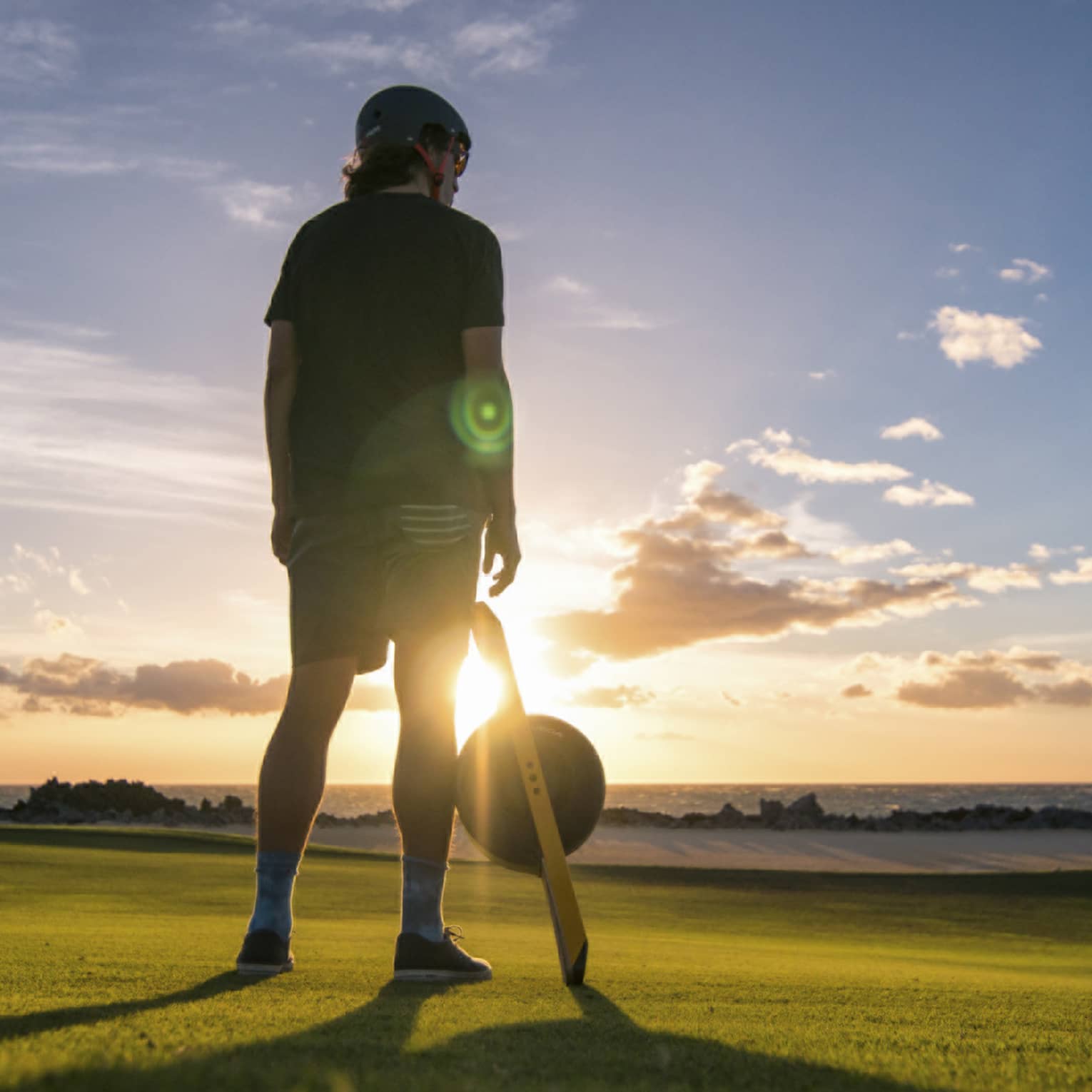 A young boy standing on a large field of green grass with the sun setting in the distance in front of him.