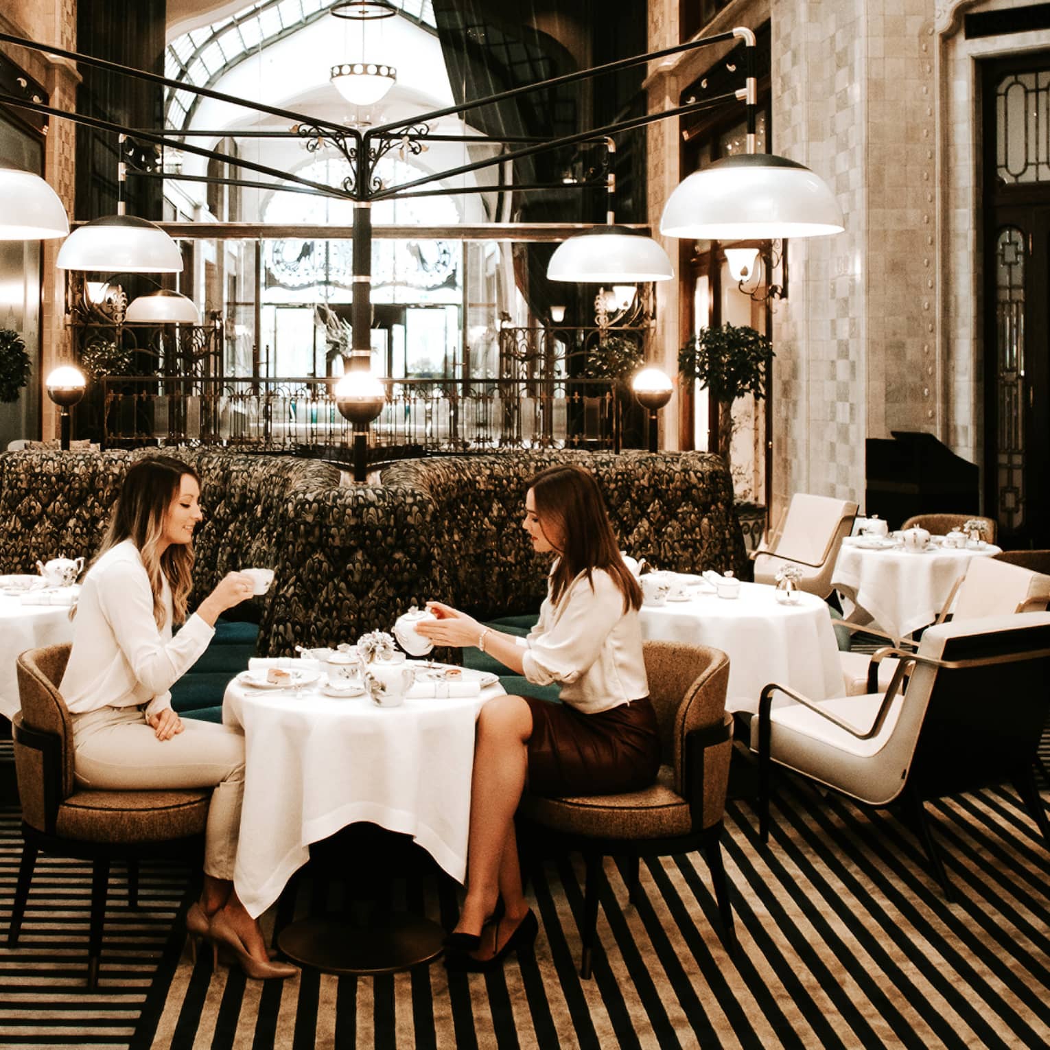Elegant dining area with a grand chandelier, high arched glass ceiling and modern seating, where two people are enjoying tea at a small table