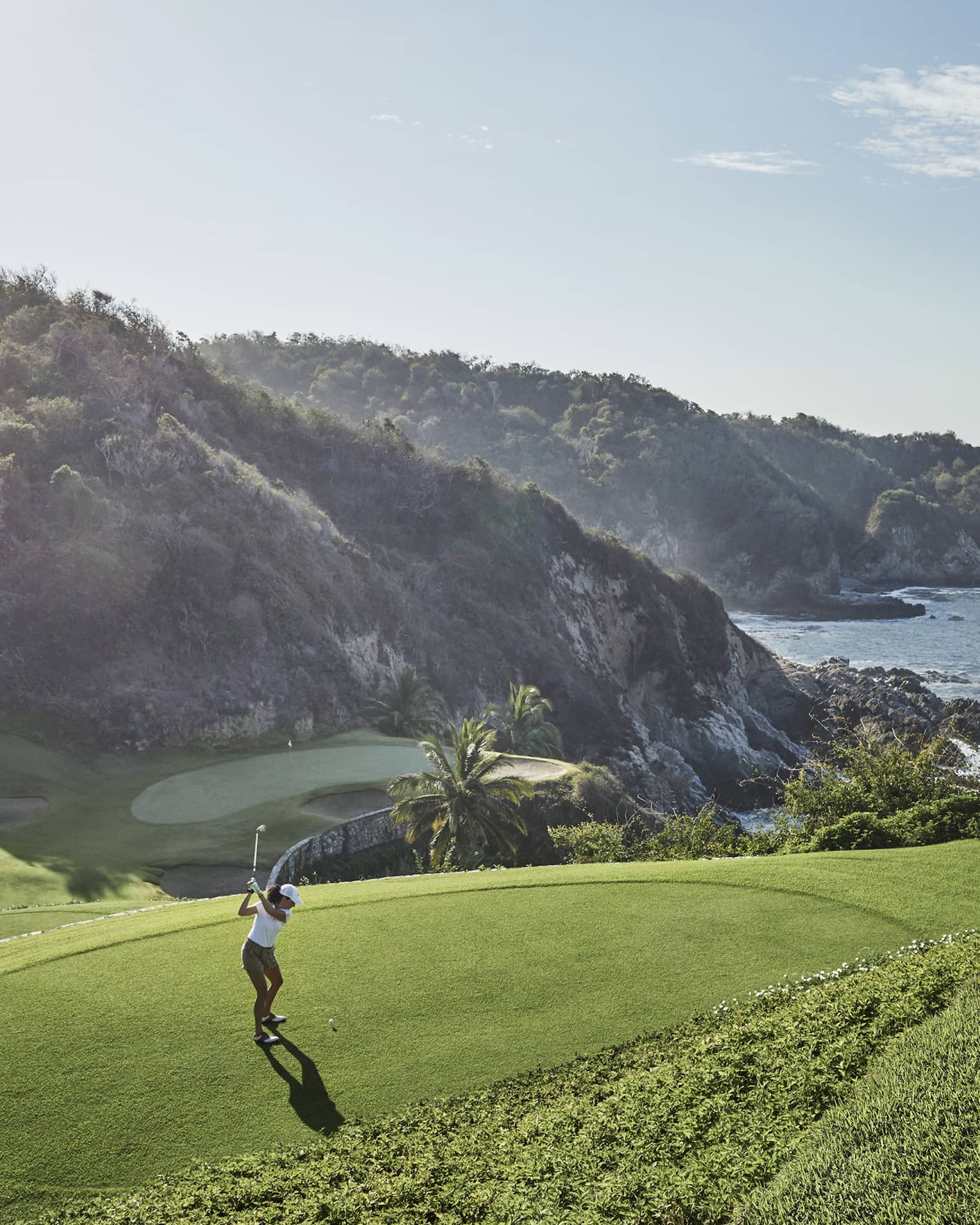 Golfer in mid-backswing, ready to strike toward a distant green at the base of an immense forested hill bordering the ocean.