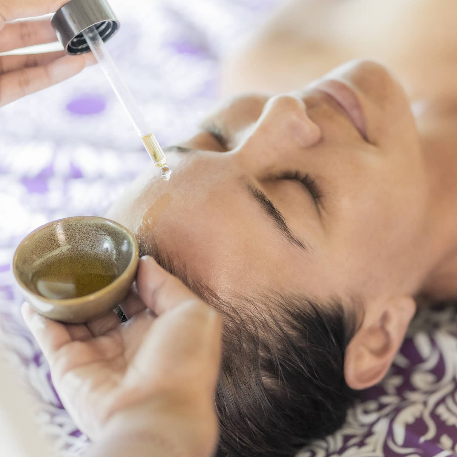 A woman receiving a relaxing facial treatment, with oil being applied from a dropper, lying on a patterned purple sheet.