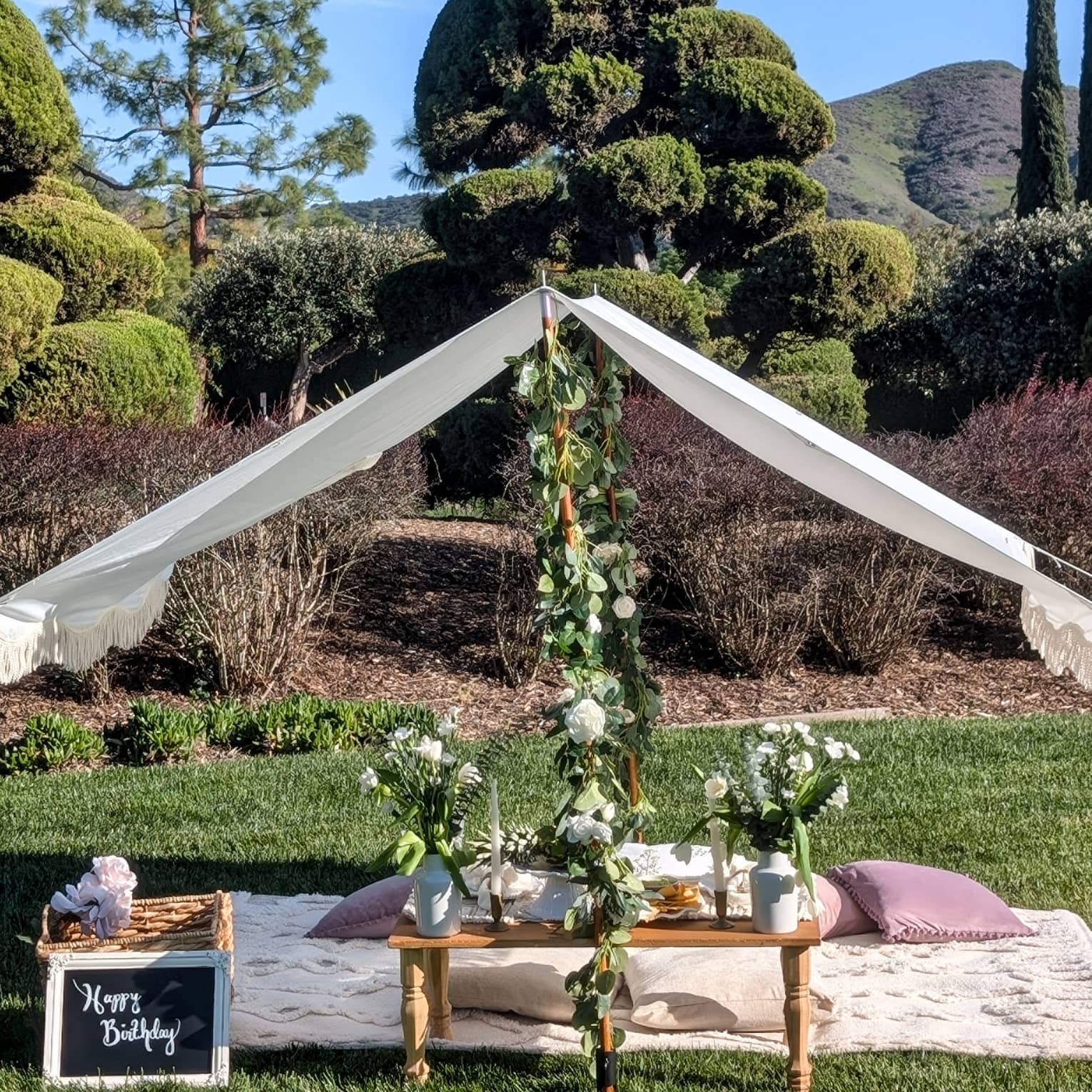 Picnic set up on a lawn with blanket, pillows, small table, flower garlands and white canopy