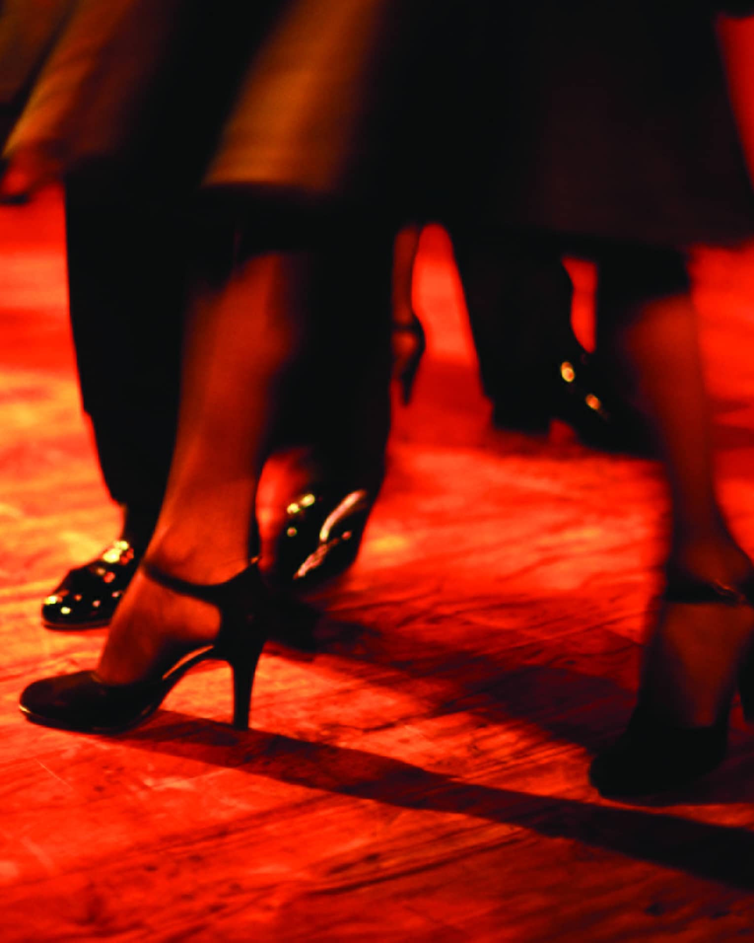 Bathed in a ruby-hued light and amidst other moving legs, a dancer's heel-clad feet step forward in a blur of motion.