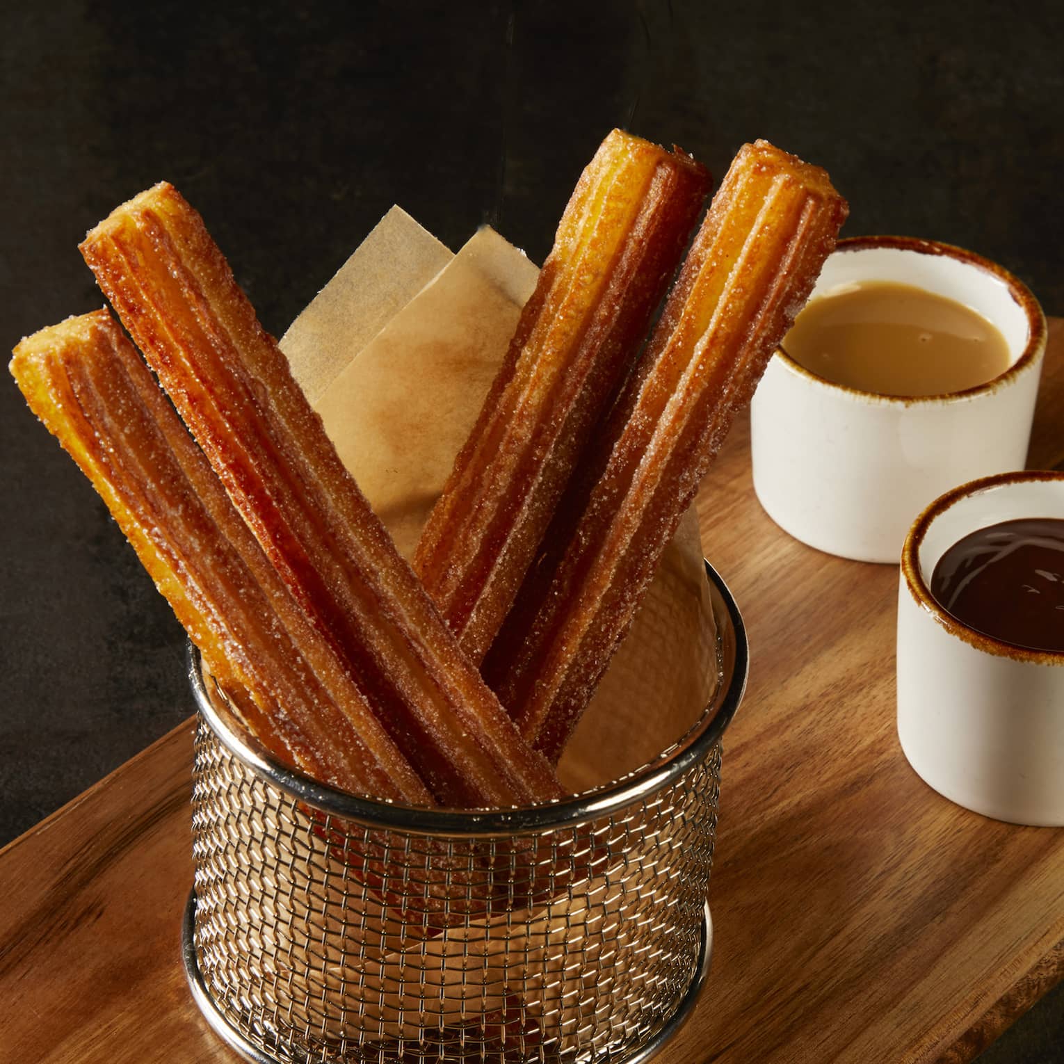 Crispy, golden churros coated in sugar in a metal basket on a wood board with chocolate and dulce de leche dipping sauces.