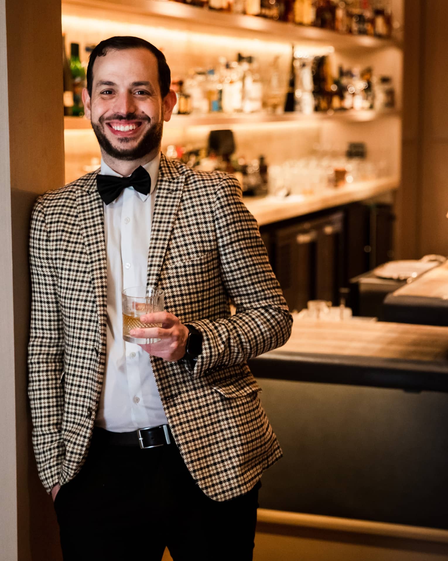 A man in a checkered suit holding a cocktail in front of a bar.