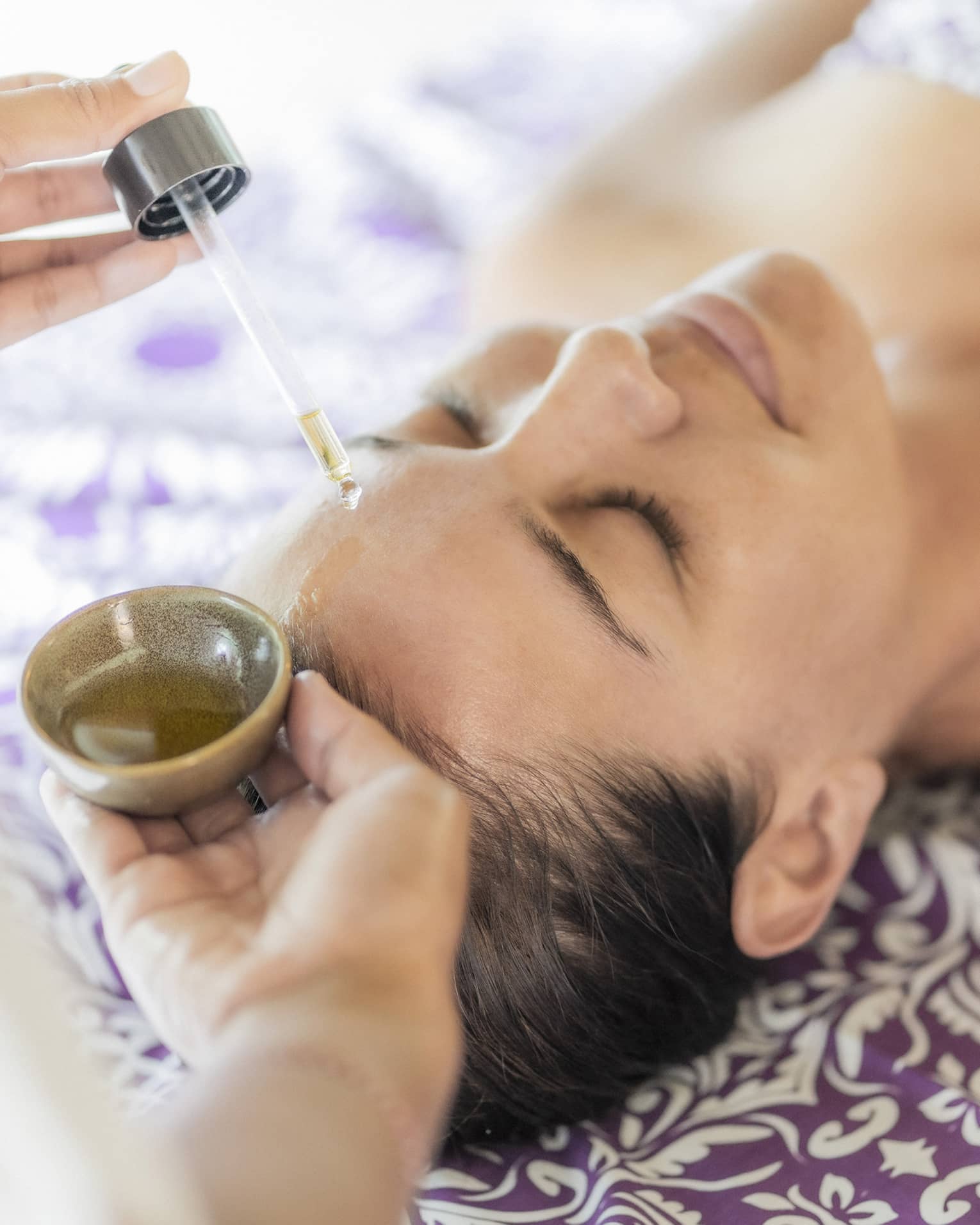 A woman receiving a relaxing facial treatment, with oil being applied from a dropper, lying on a patterned purple sheet.