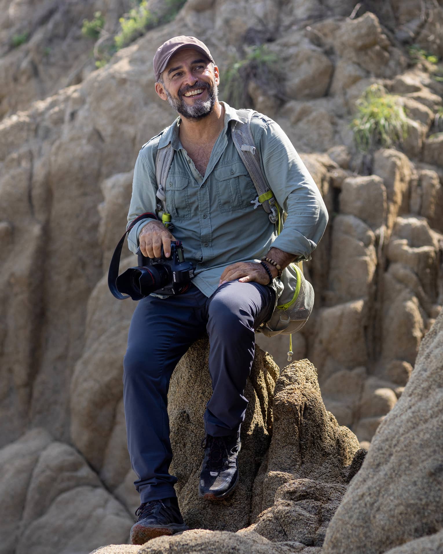 A photographer wearing a backpack and holding a camera in one hand sits atop a rock formation, smiling into the distance.