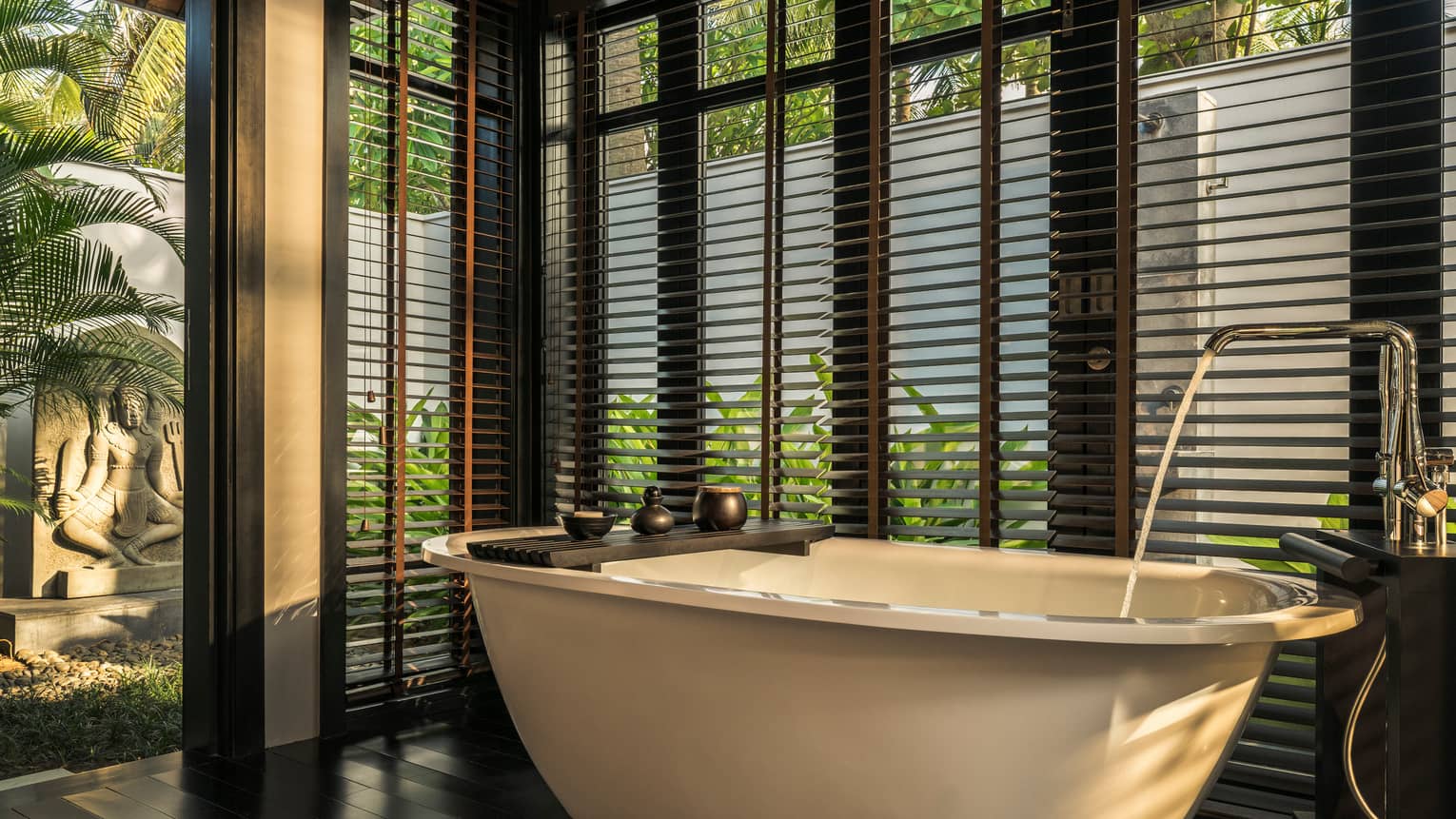 Luxurious bathroom with a free-standing bathtub, surrounded by large windows with wooden blinds, overlooking a garden with a stone sculpture and tropical plants.