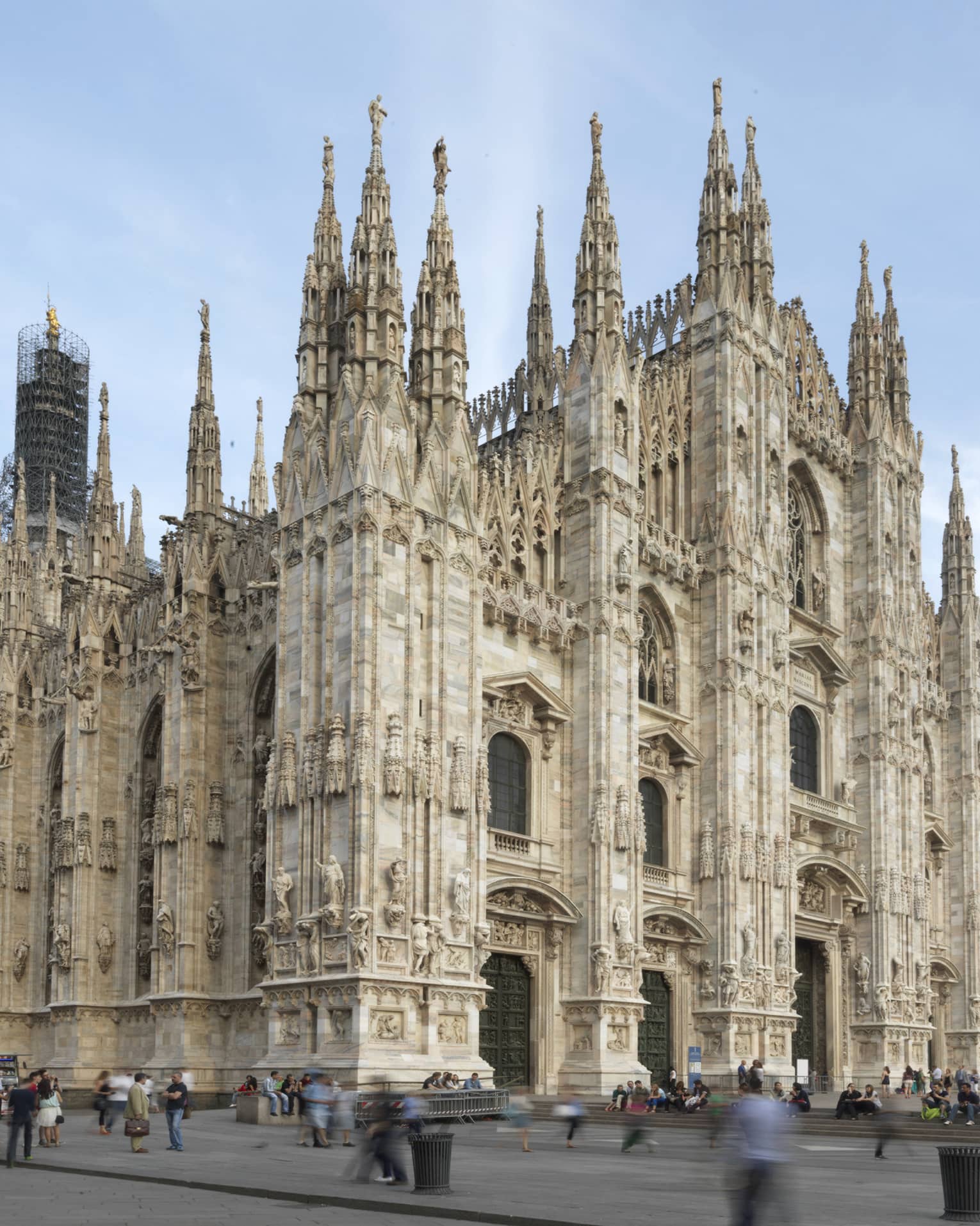 The exterior of the Milan Cathedral (Duomo di Milano), showcasing its intricate Gothic architecture with tall spires and detailed stone carvings against a clear sky.