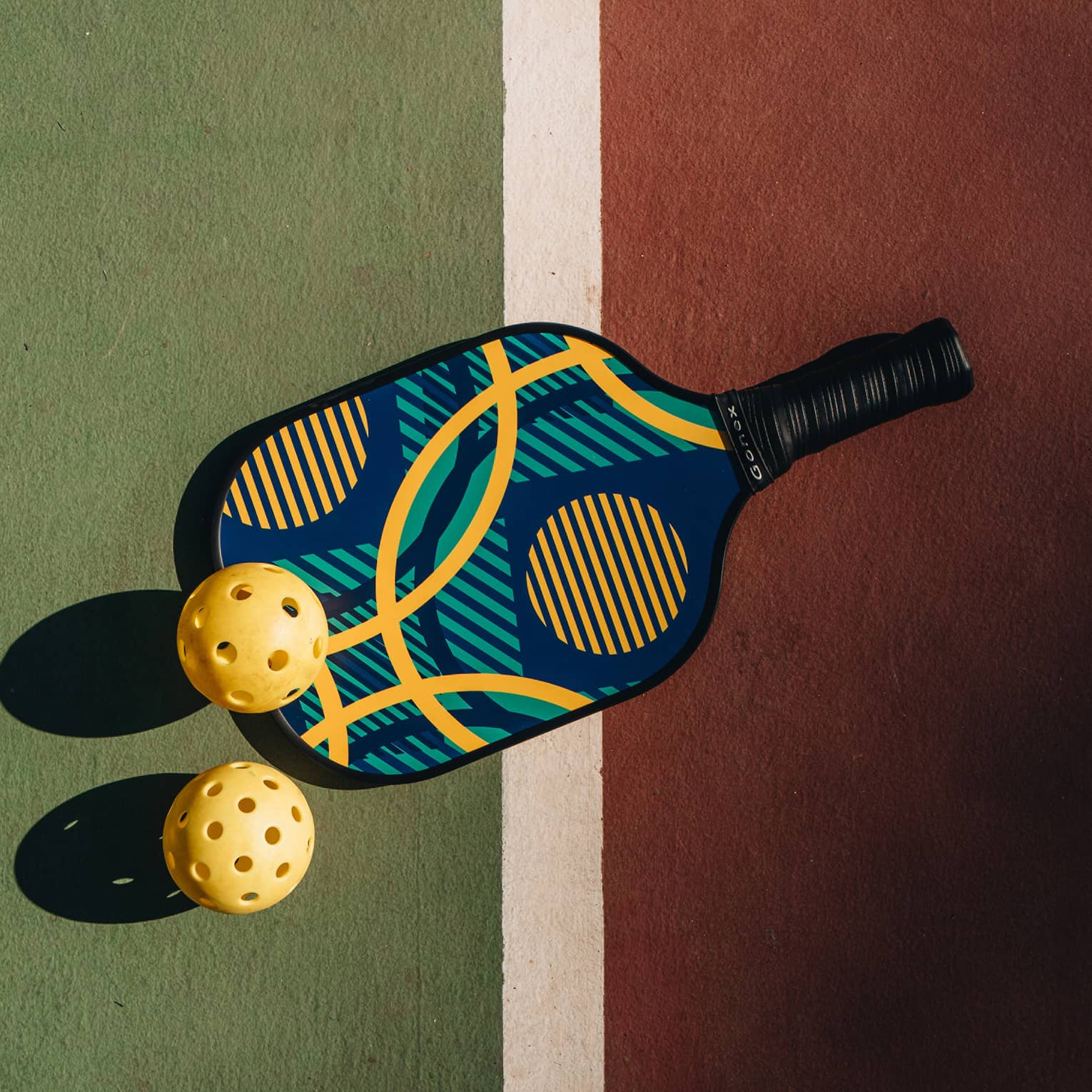 A colourful pickleball racquet and two yellow pickleballs cast shadows upon a green and red court with a white dividing line.