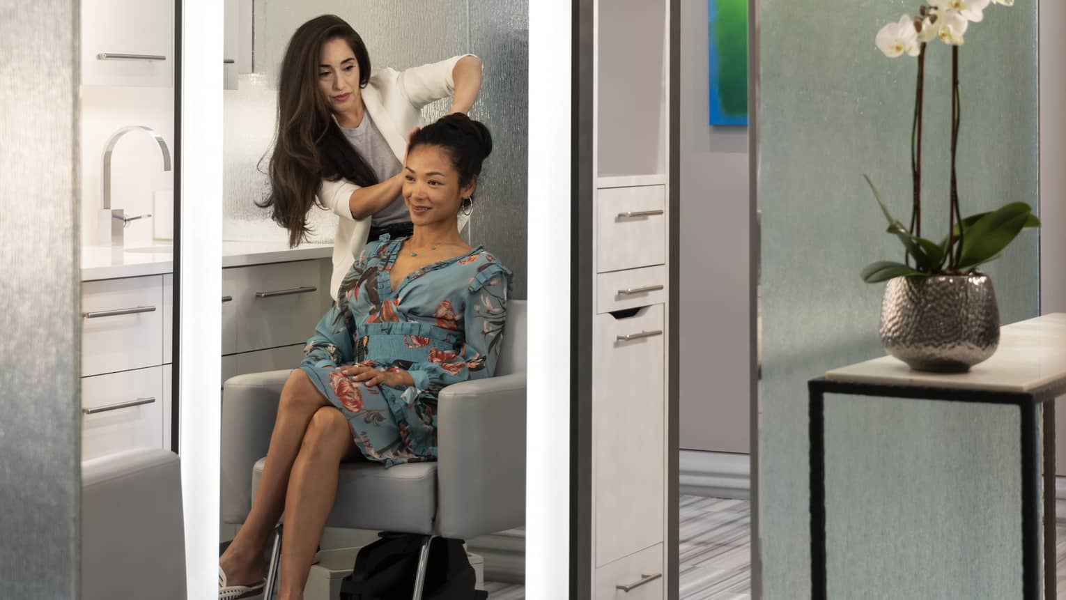 A woman styles another woman's hair; both reflections are seen in an illuminated mirror at salon