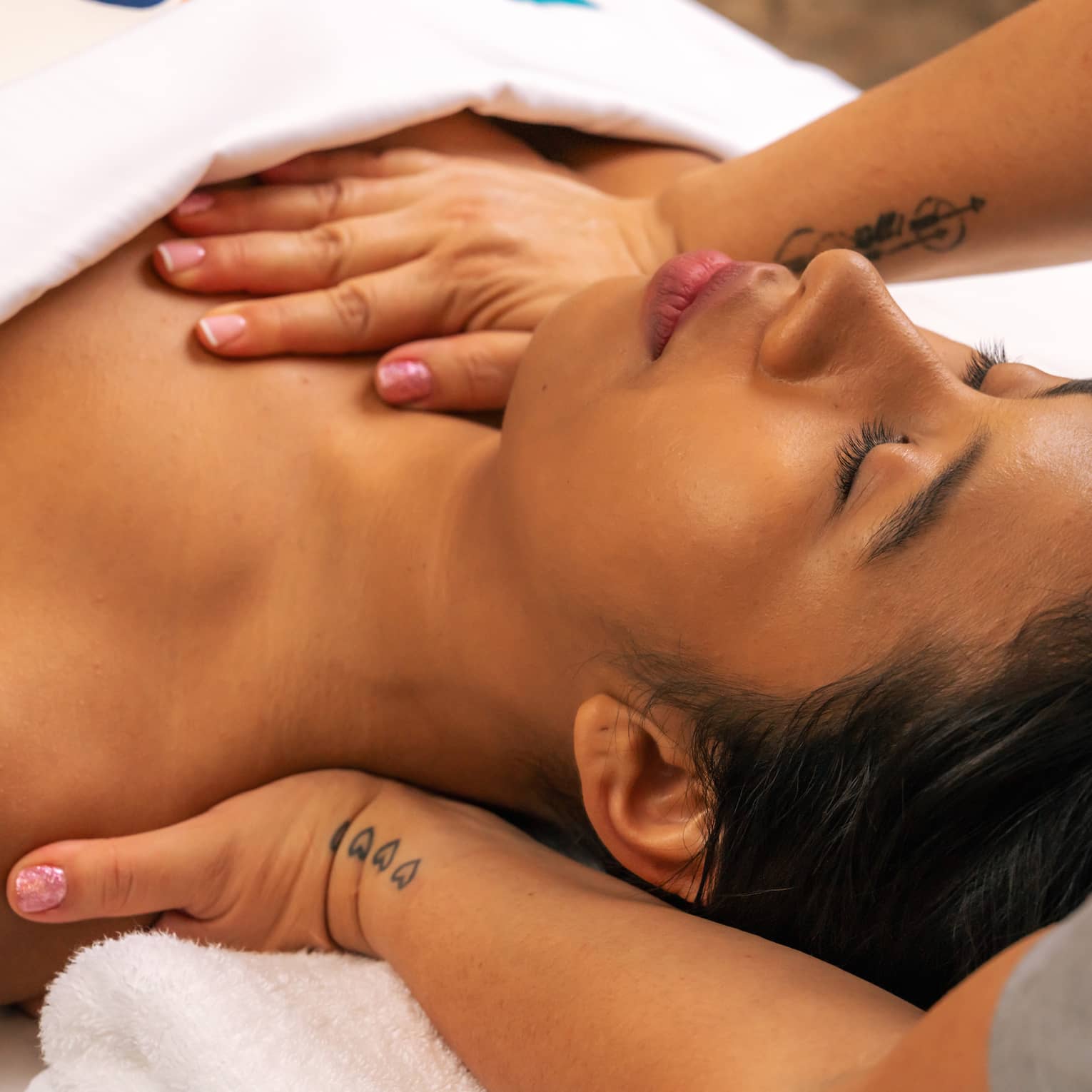 Person lies on their back beneath a white blanket with their eyes closed as a pair of hands massages the back of their shoulder and applies pressure on sternum