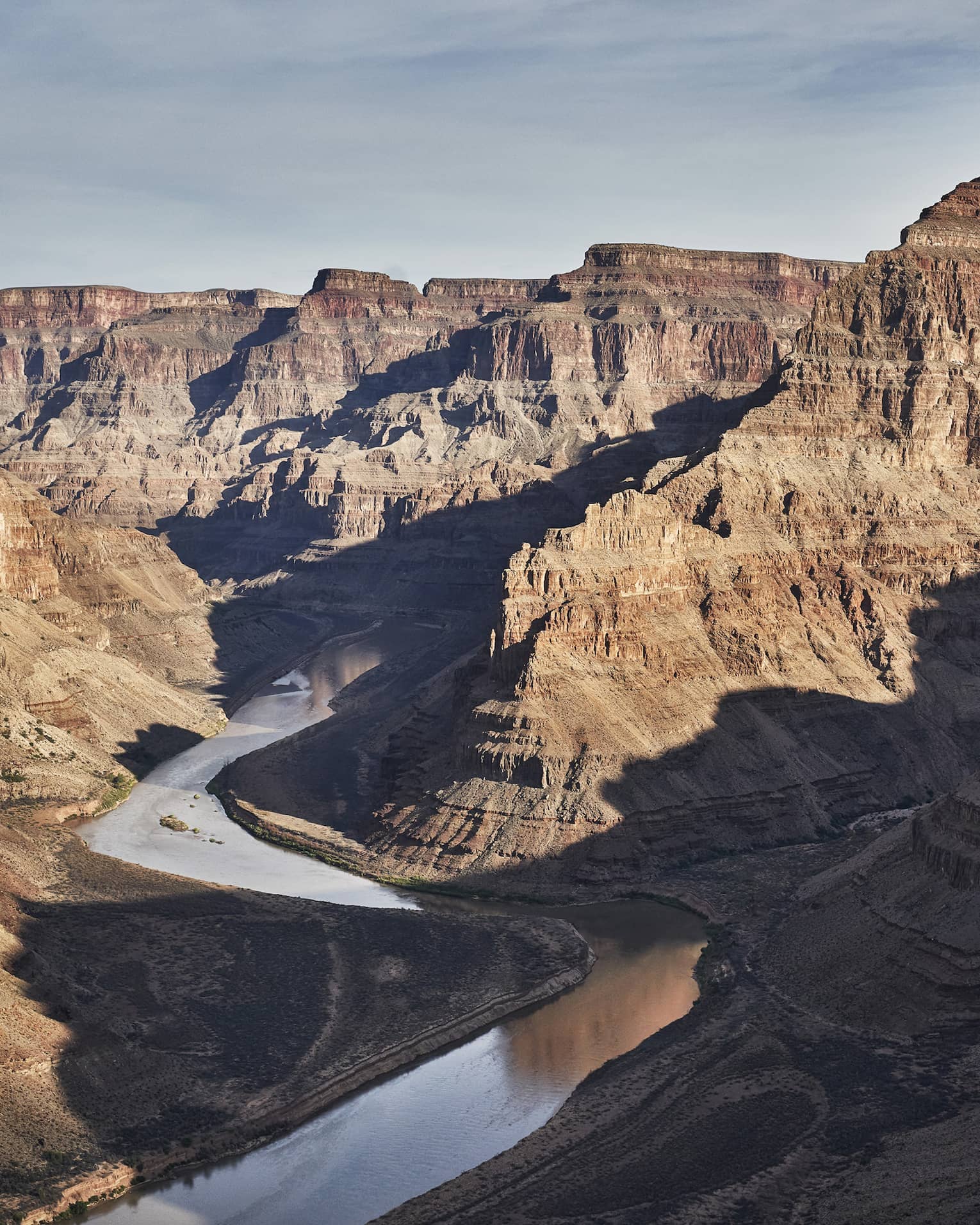 A silvery river meanders its way along the bottom of a wide, deep canyon with flat-topped ridges of rock towering upwards.