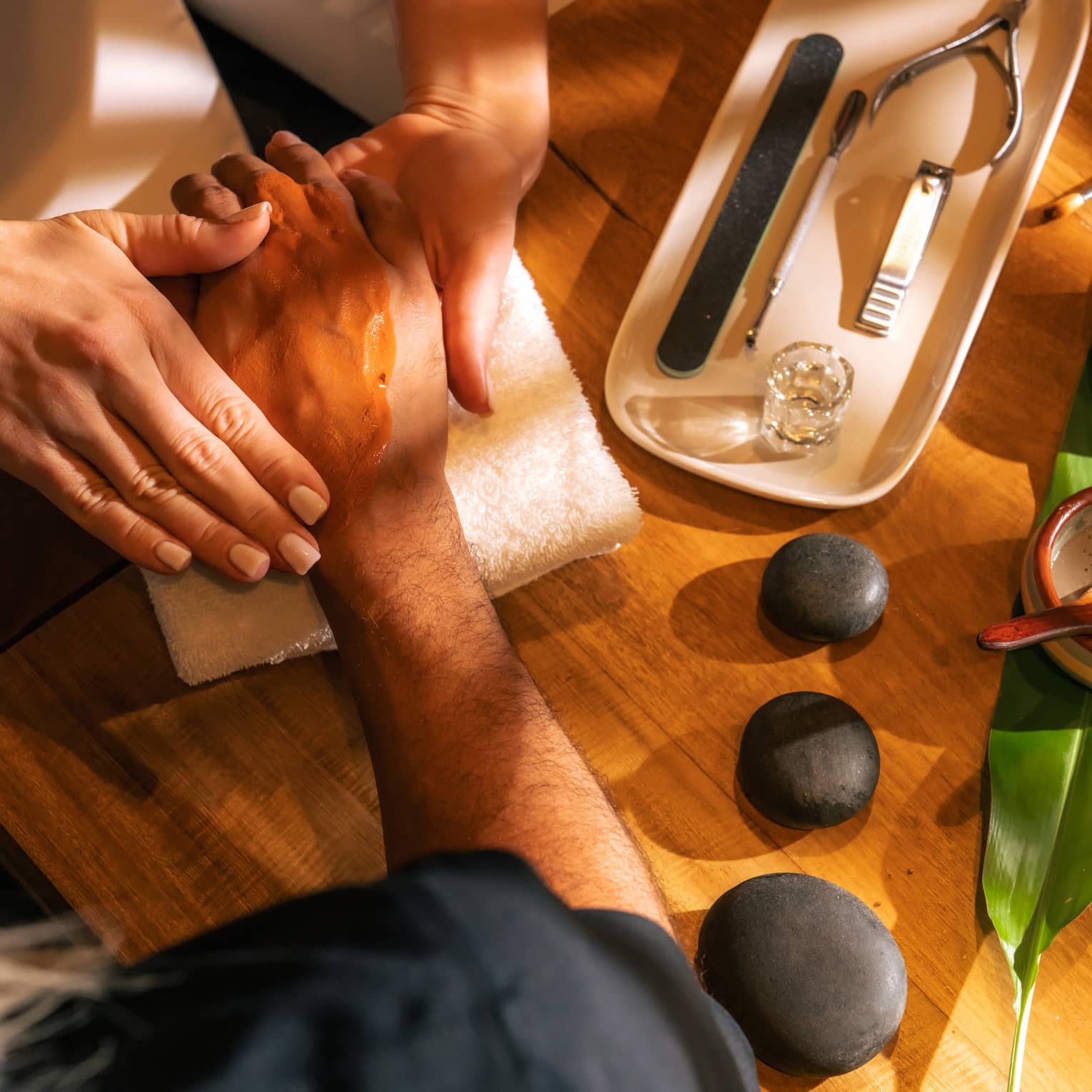 Manicurist massages a clients hand with three black stones, two small bowls of ingredients and nail tools sitting on the table