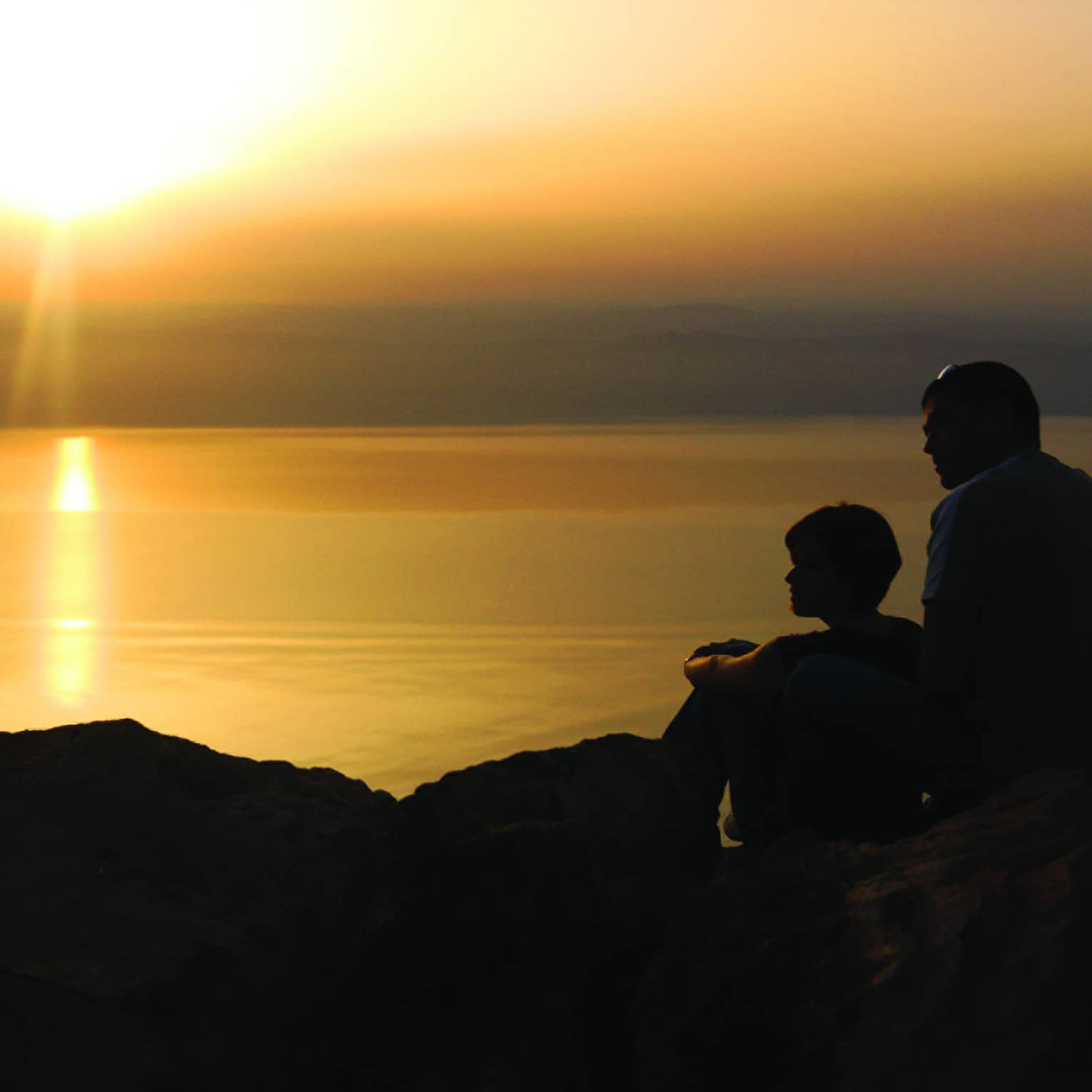 A couple sitting on a rocky outcrop are silhouetted against a setting sun that casts an orange glow on the lake before them.
