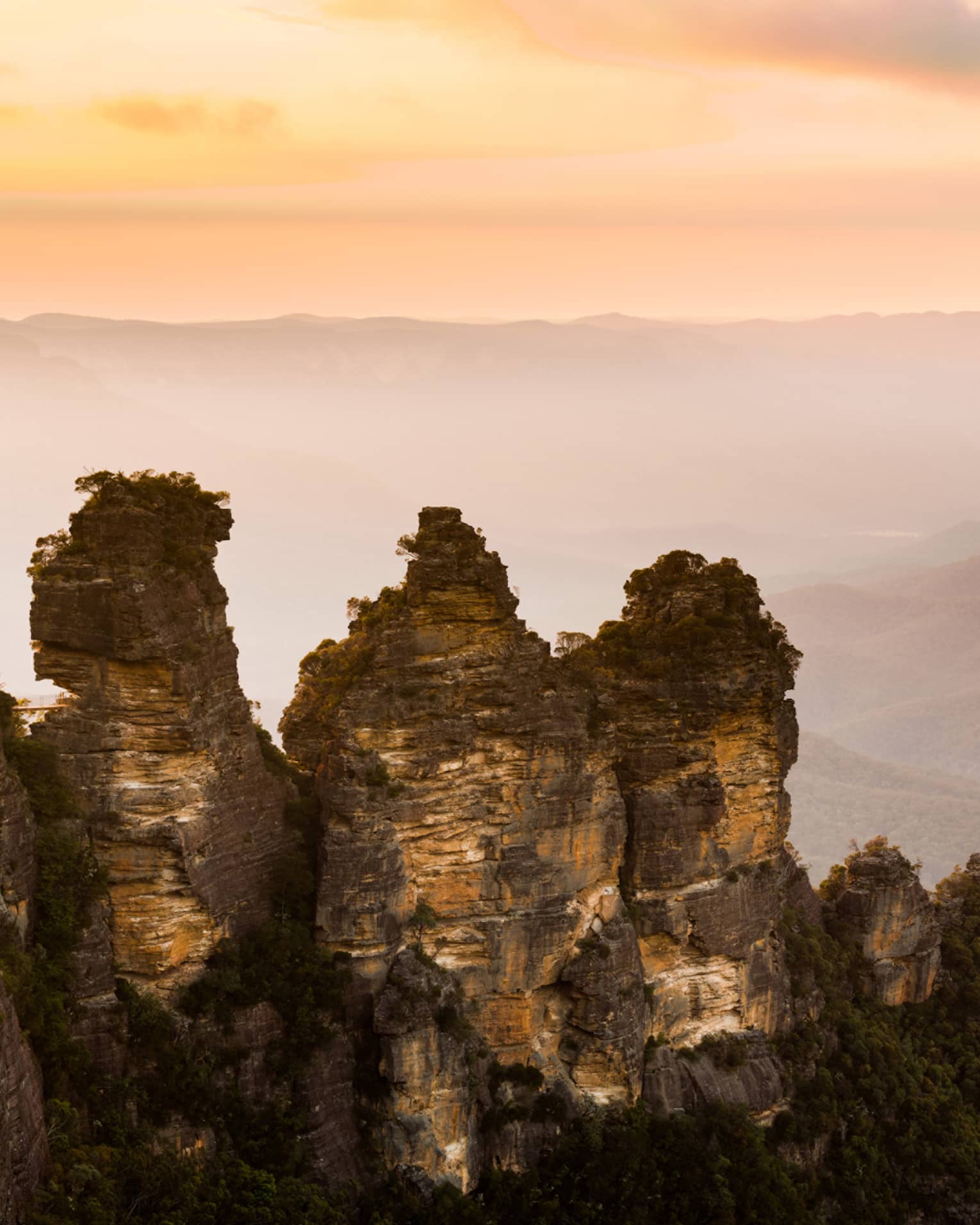 Dramatic sunset view looking out across misty mountains, a wall of craggy, sparsely treed rocks rising up in the foreground.