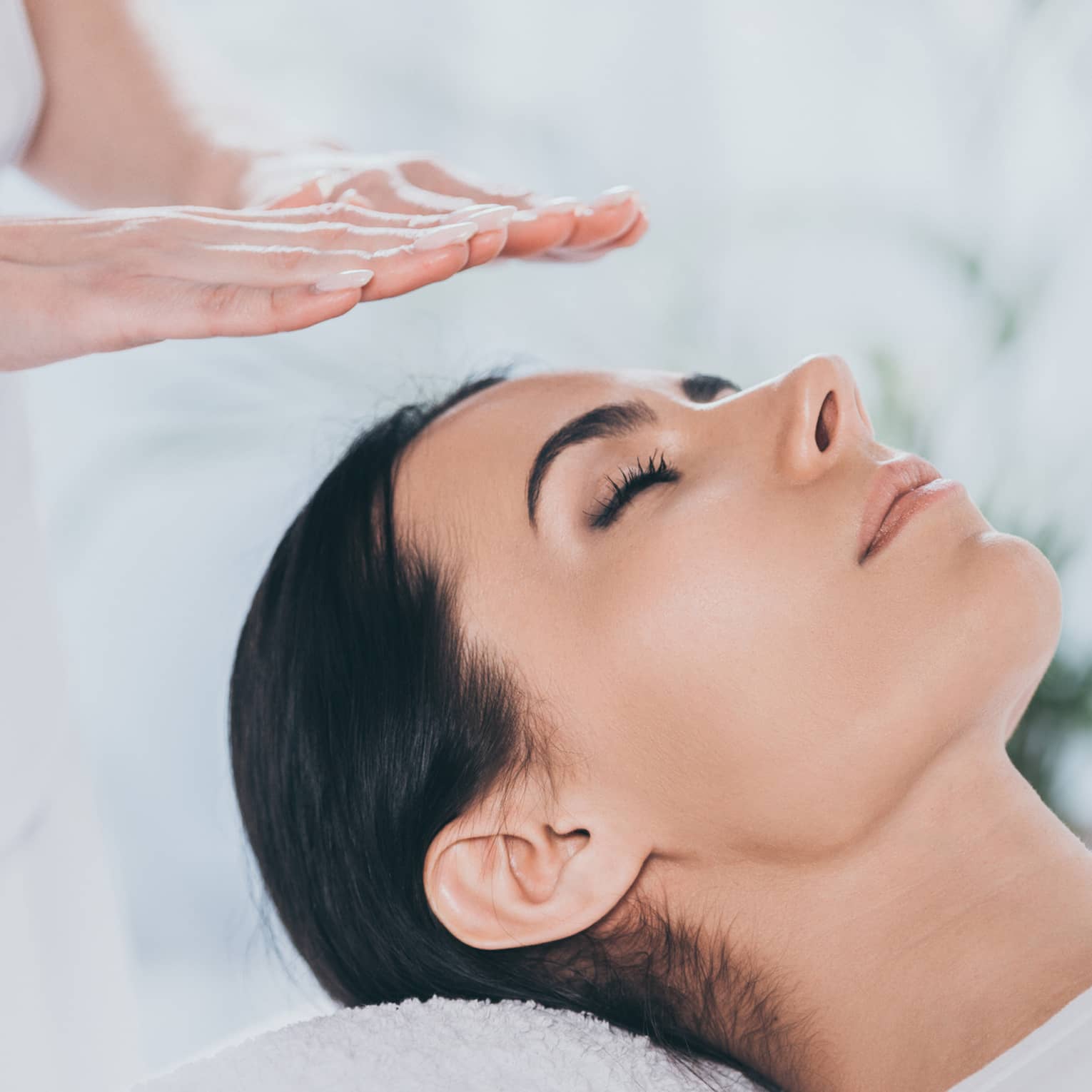 Woman with eyes closed lays on rolled up towel on spa table under therapists' hovering hands