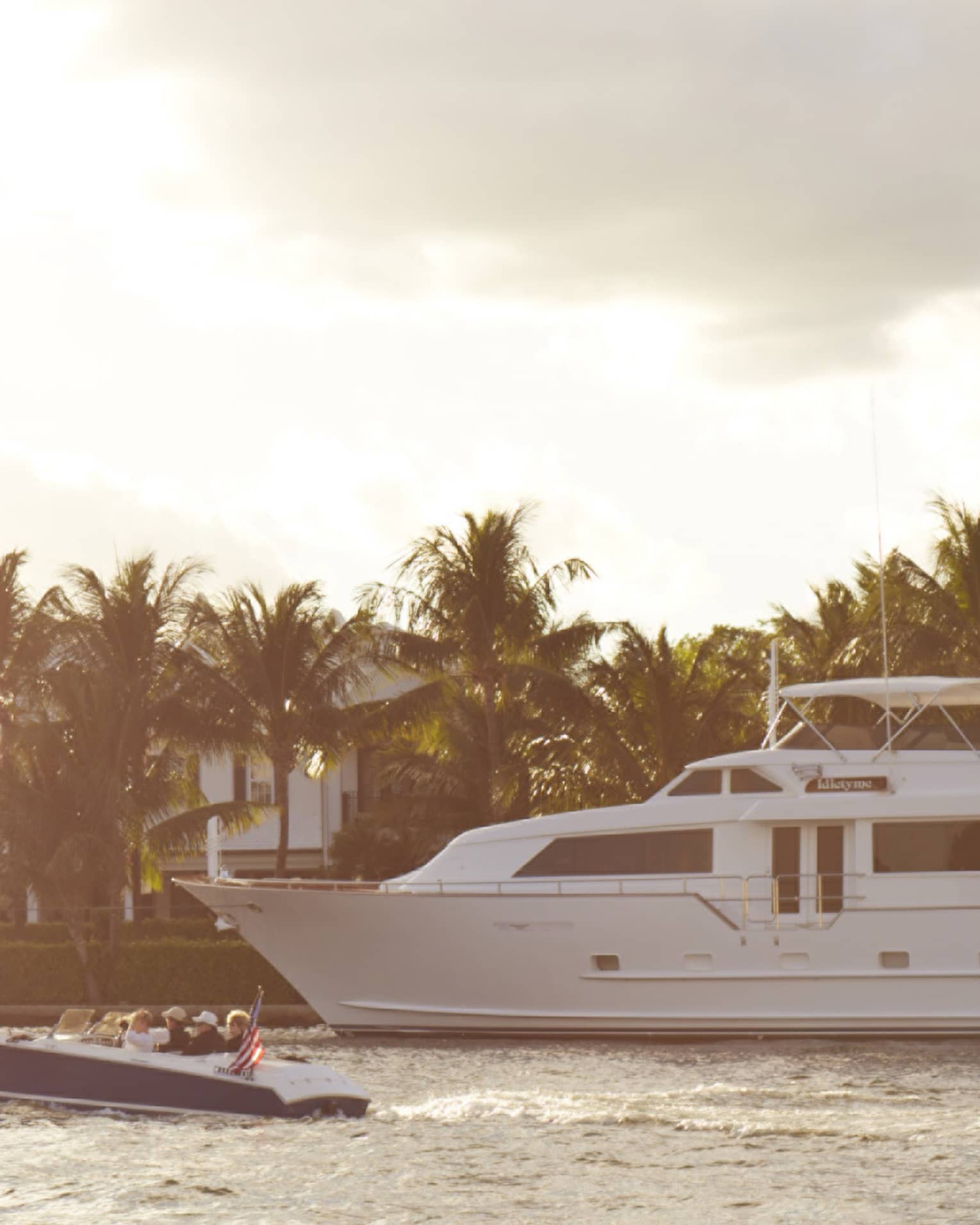 A large white yacht cruises alongside a smaller boat in a serene waterway, with palm trees and luxurious homes in the background under a partly cloudy sky.