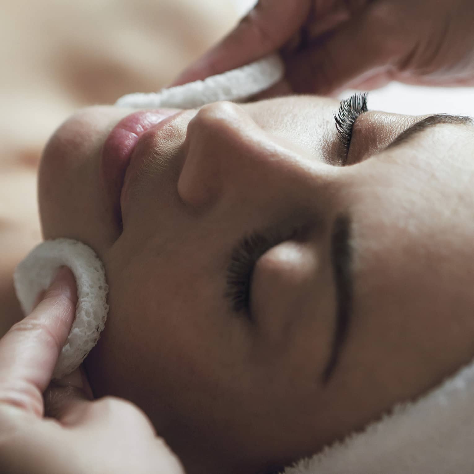 A detail of hands rubbing a sponge on a woman's face as she lies on a massage table with her eyes closed and hair wrapped up in a white towel