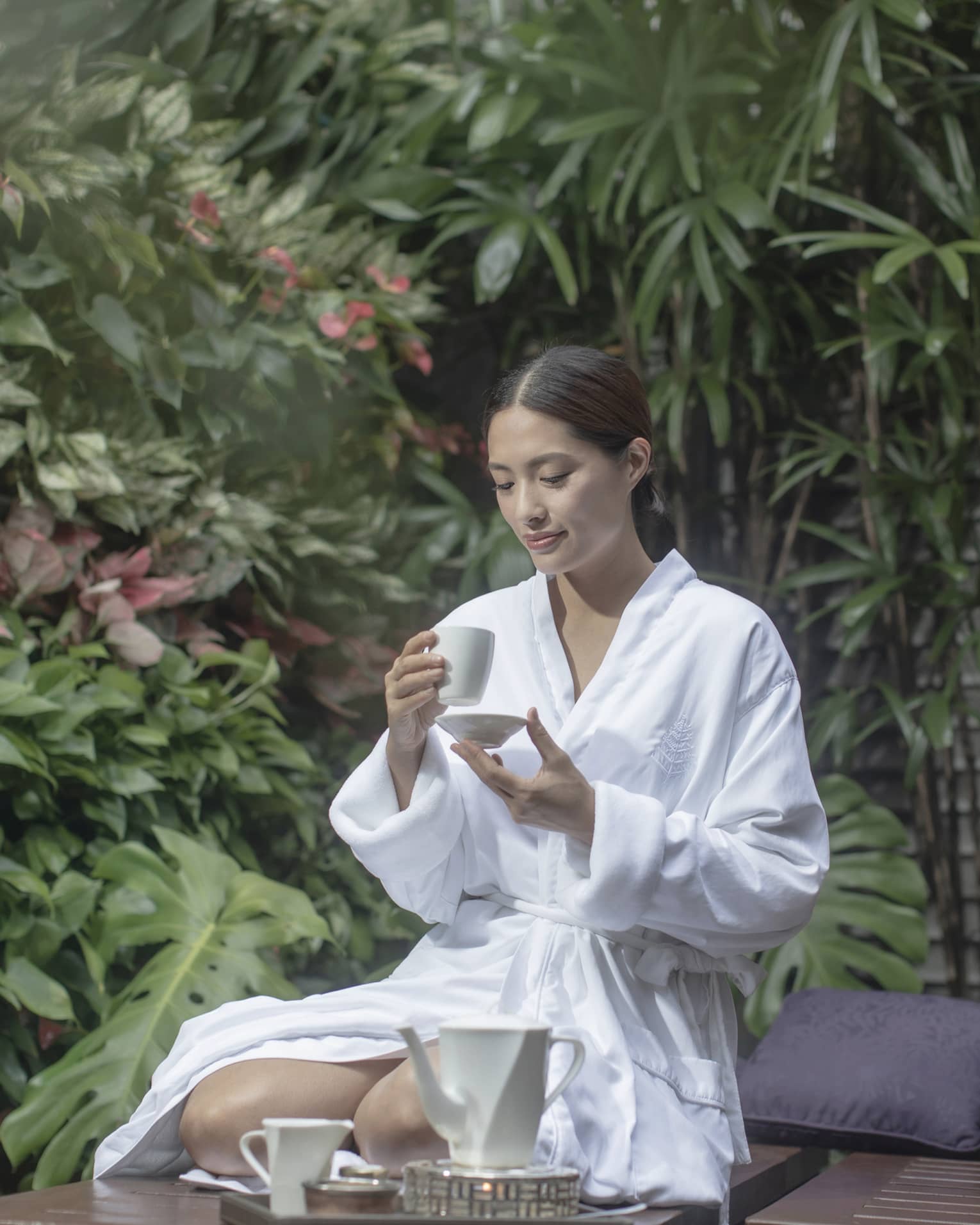 Woman in white spa robe kneeling and sipping tea among outdoor foliage