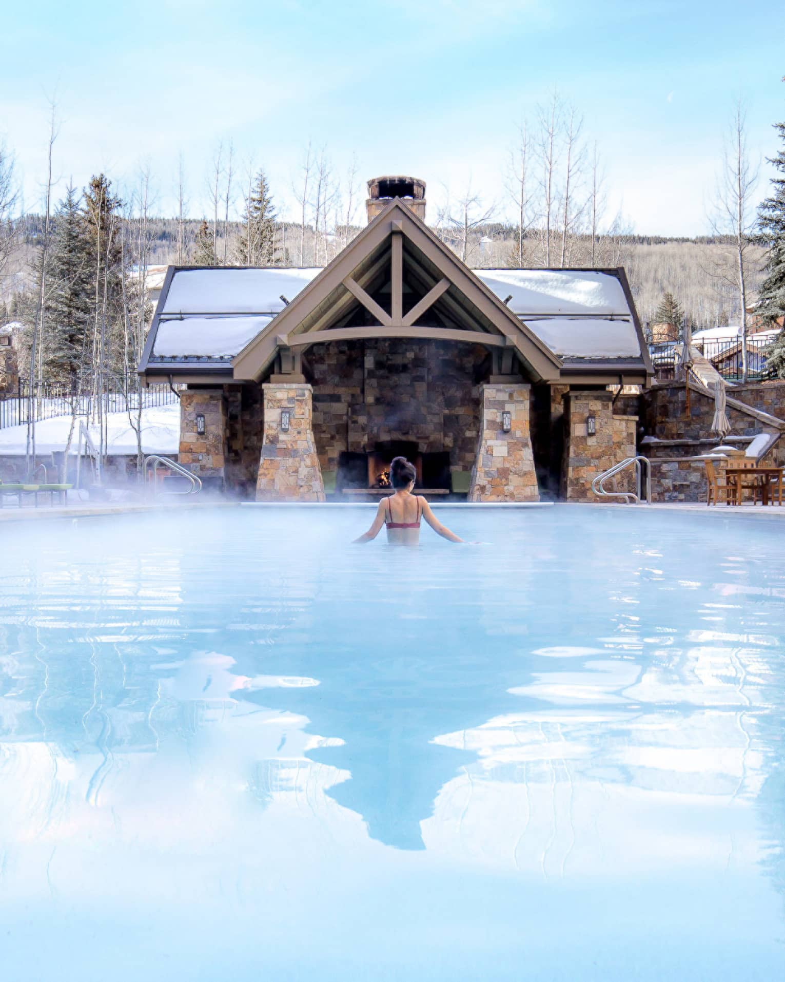 A woman in a pool with a cabin in front of her and snow covered trees around her.