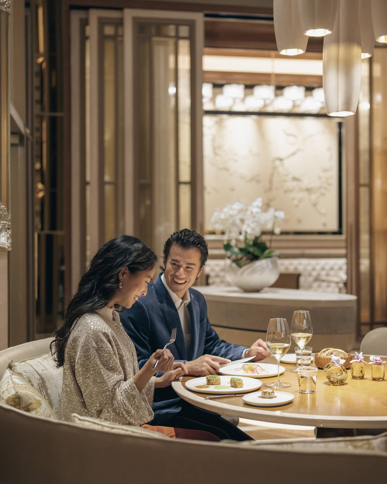 A smiling couple savours lunch with wine in a circular booth at a fine dining restaurant with warm pendant lighting.