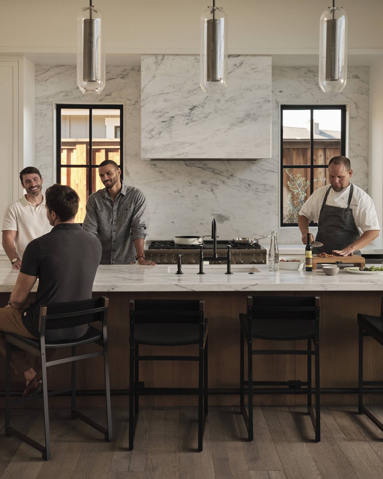 A group of guests gather at one end of a long kitchen island while a chef chops ingredients at the other end of the island.