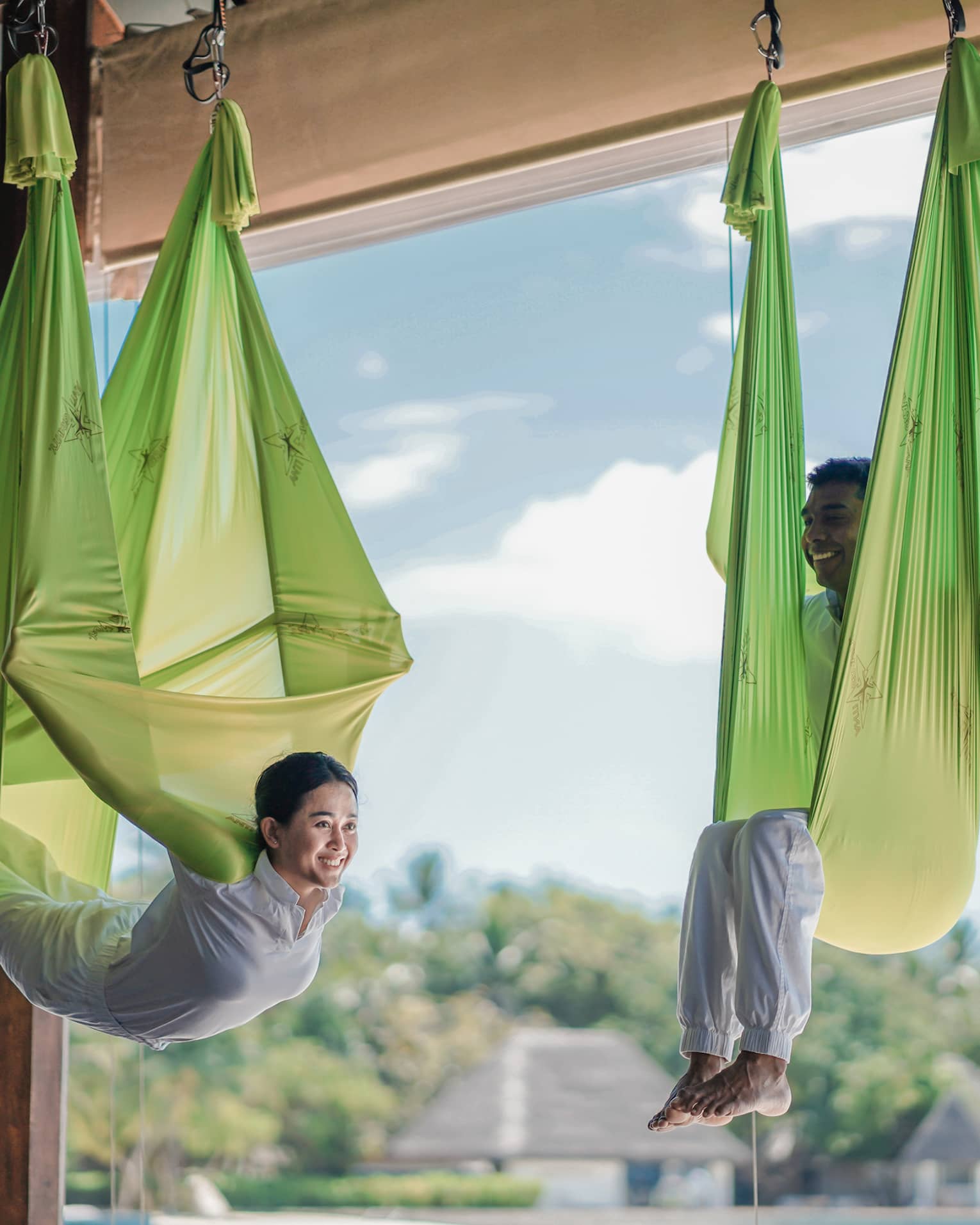 Two people practising antigravity yoga in bright green hammocks, suspended in a wellness resort studio with a view of lush tropical surroundings