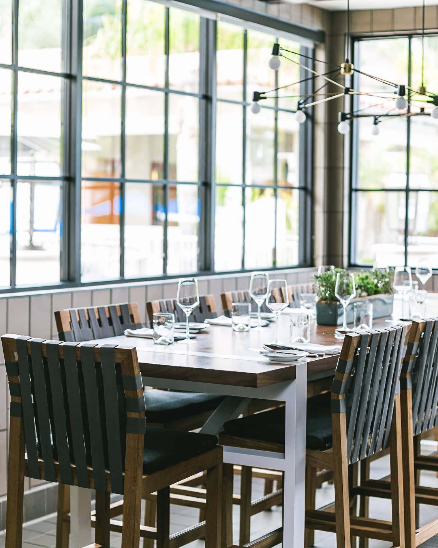 A rectangular wooden dining table with 12 black chairs under an industrial chandelier in a bright room with tiled walls.
