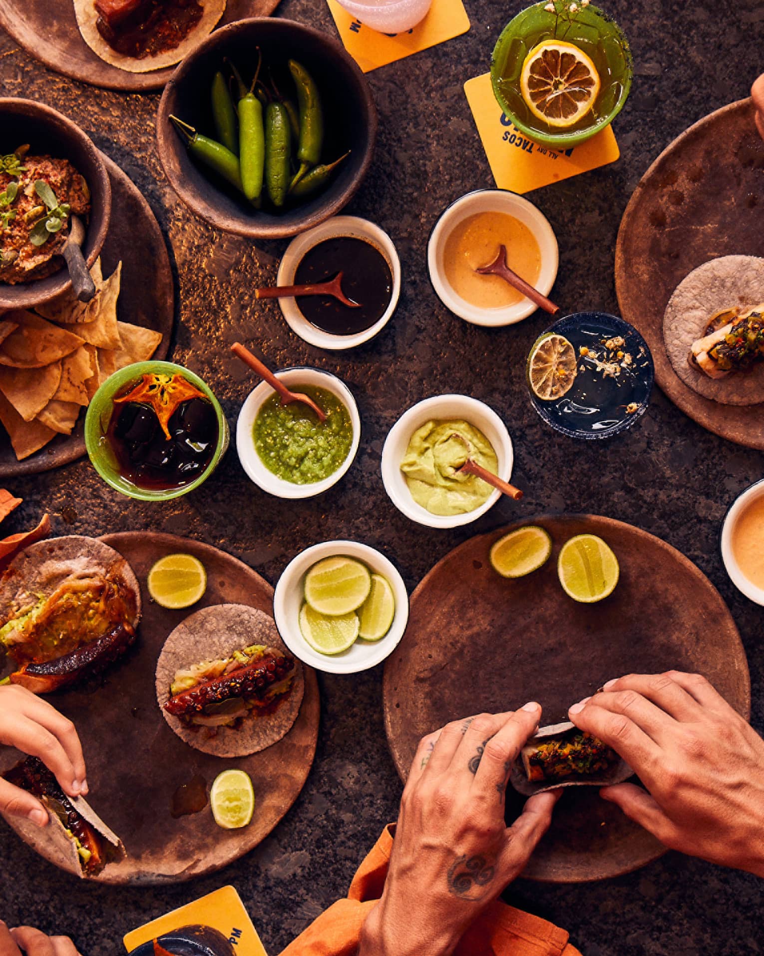 An overhead view of five pairs of hands holding tacos next to bowls of chips, salsa, peppers, limes and sauces.