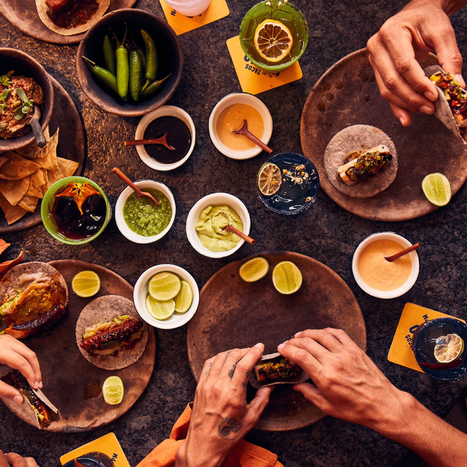 An overhead view of five pairs of hands holding tacos next to bowls of chips, salsa, peppers, limes and sauces.