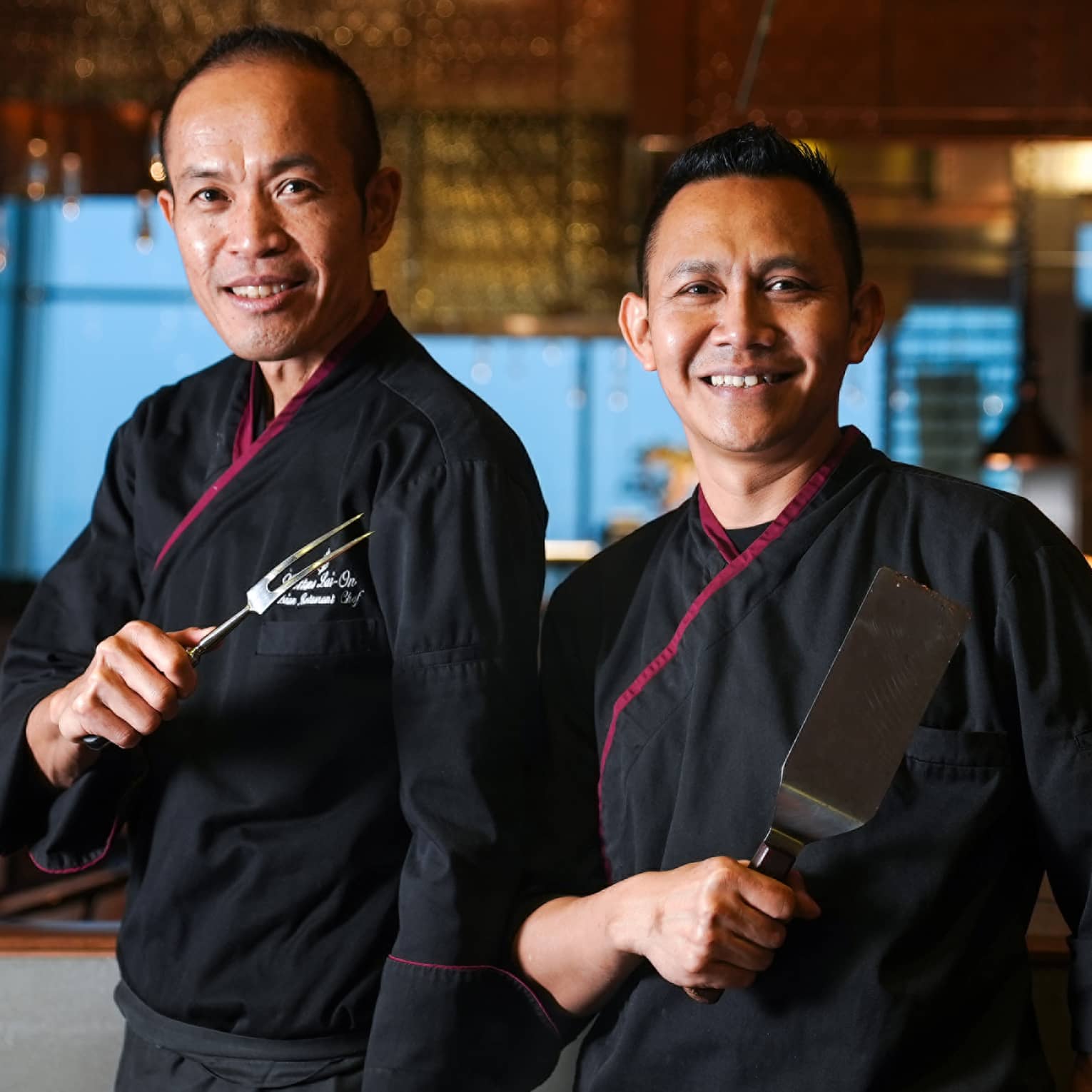 Two smiling chefs, dressed in black chef jackets and holding cooking utensils, pose together in front of the dining area.