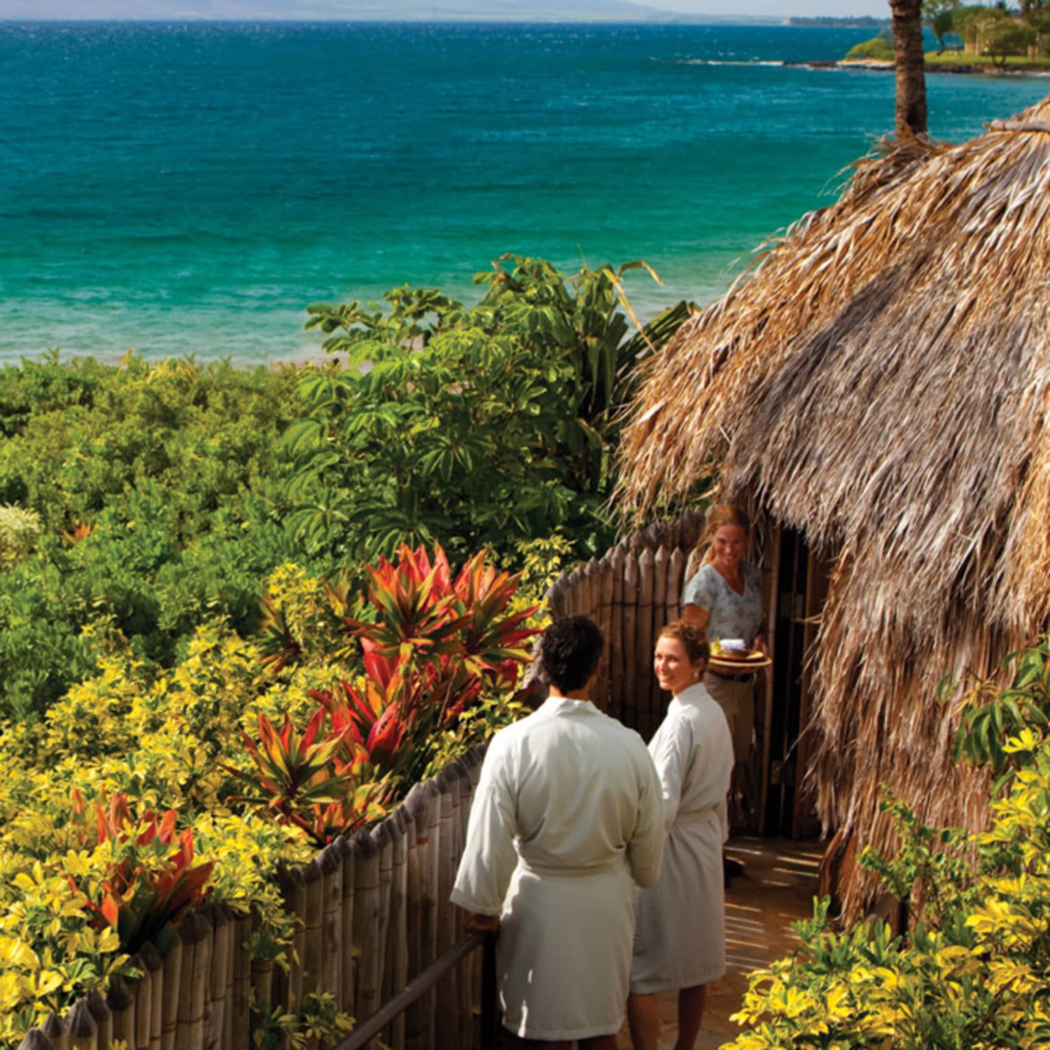 Couple in white bathrobes follow spa attendant into thatched-roof hut on path looking out at ocean