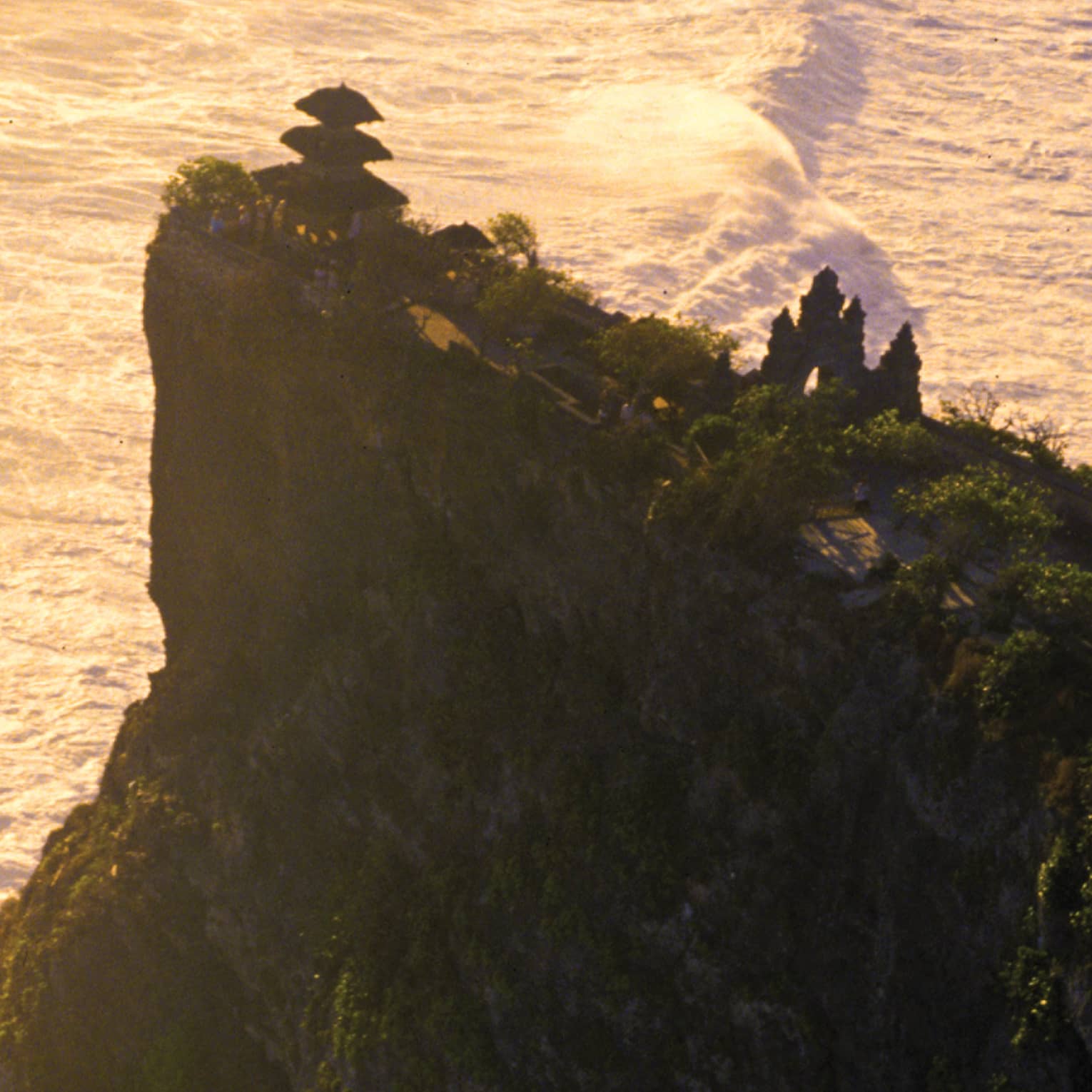 Aerial view of a temple nestled among trees at the edge of a tall, steep cliff; big, white waves crash in the vast sea below.