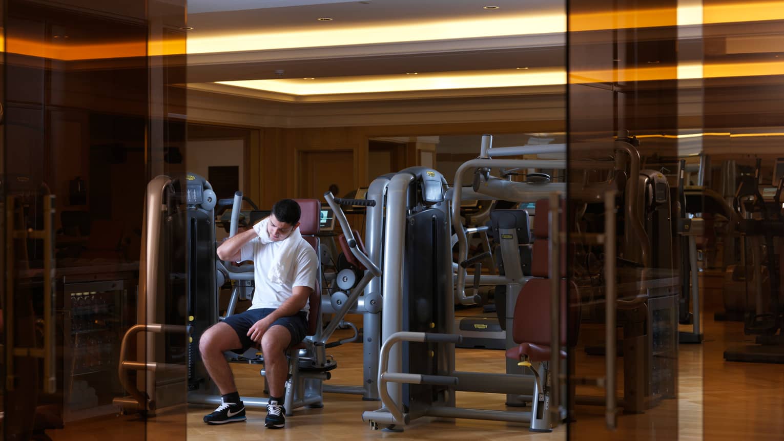 Man sits on weights machine, white towel around neck, past orange glass doors in fitness centre