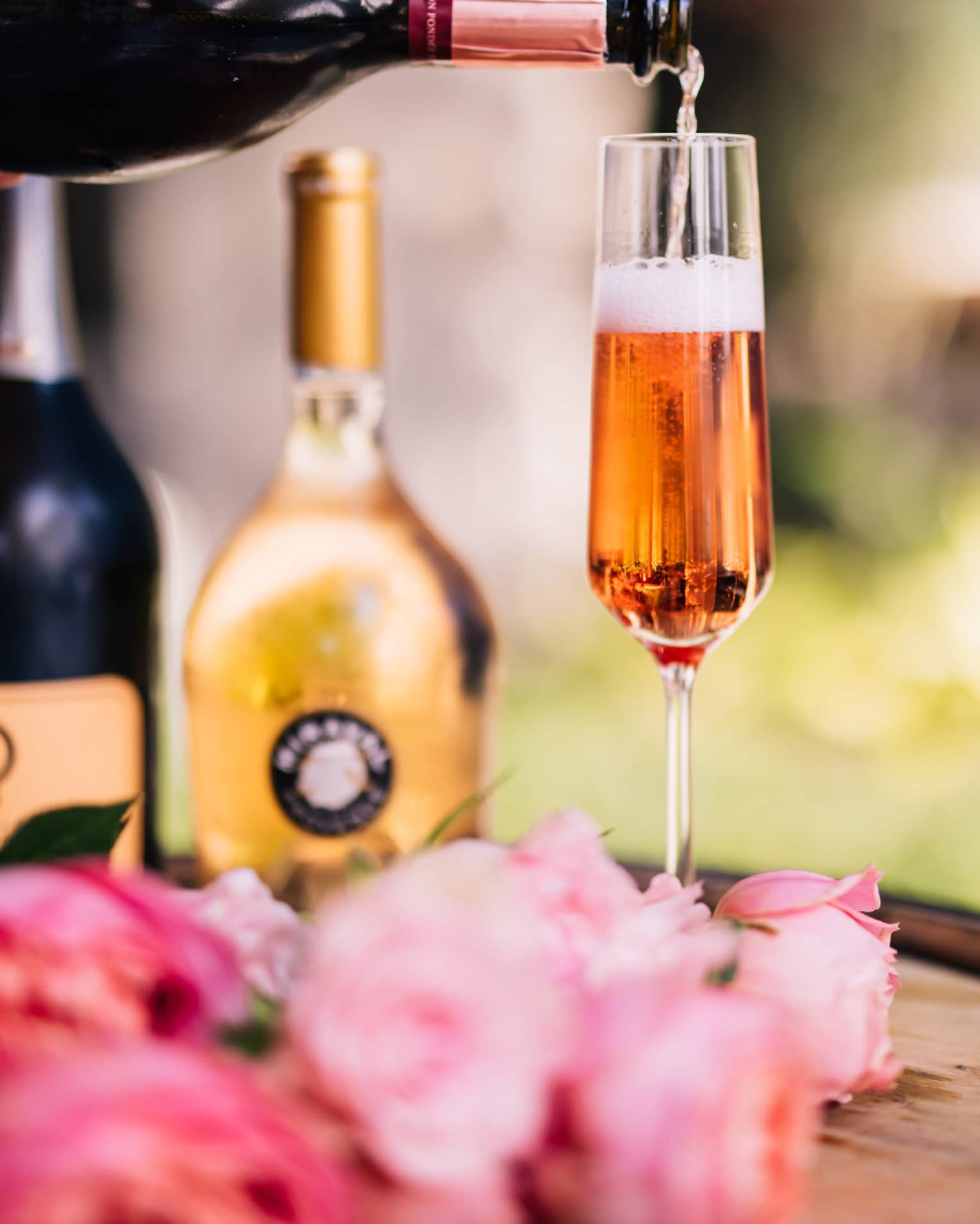 A glass of rose next to rose petals and bottles.