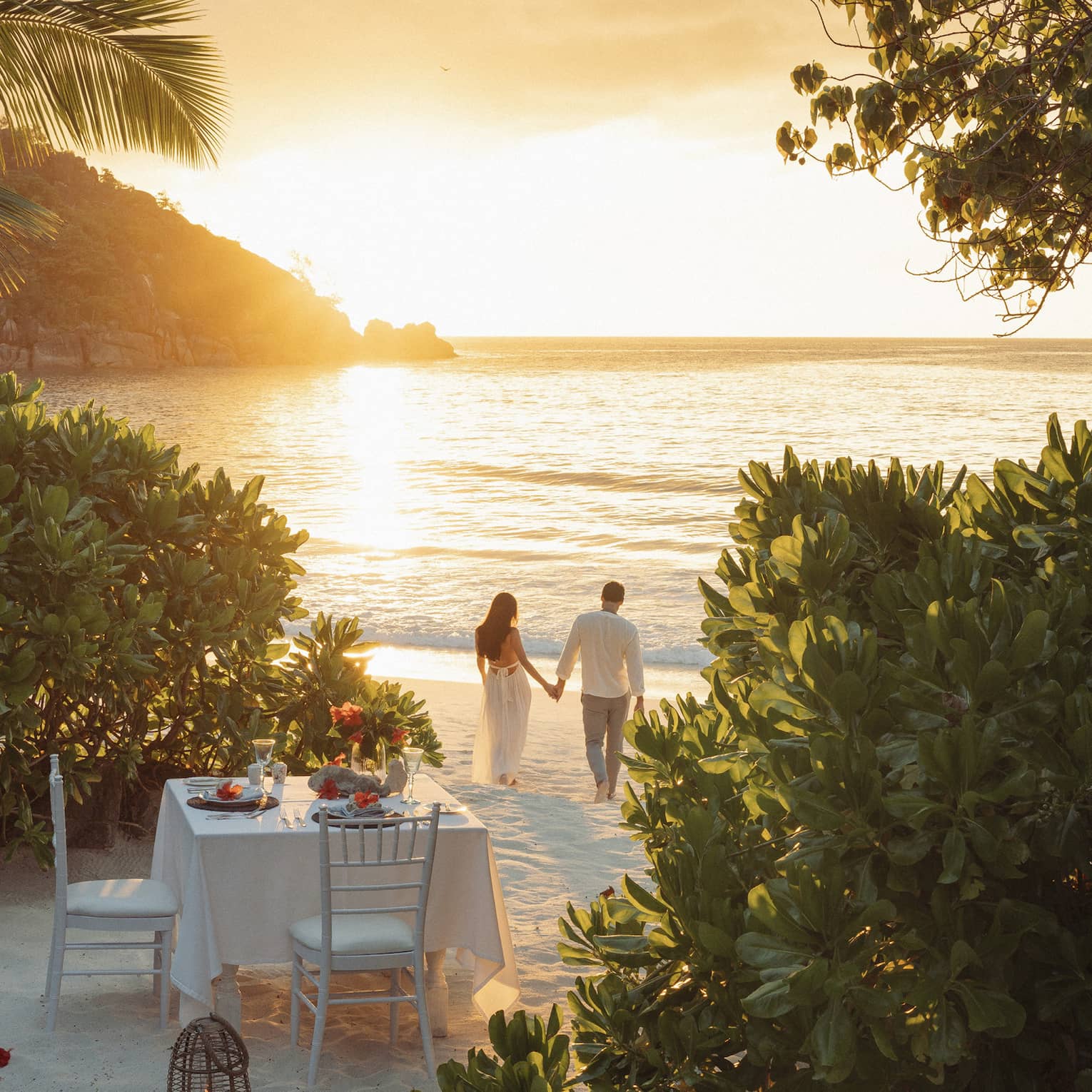 Framed by trees, a romantic dinner table set for two on the beach; a couple holding hands walks in the golden glow of sunset.