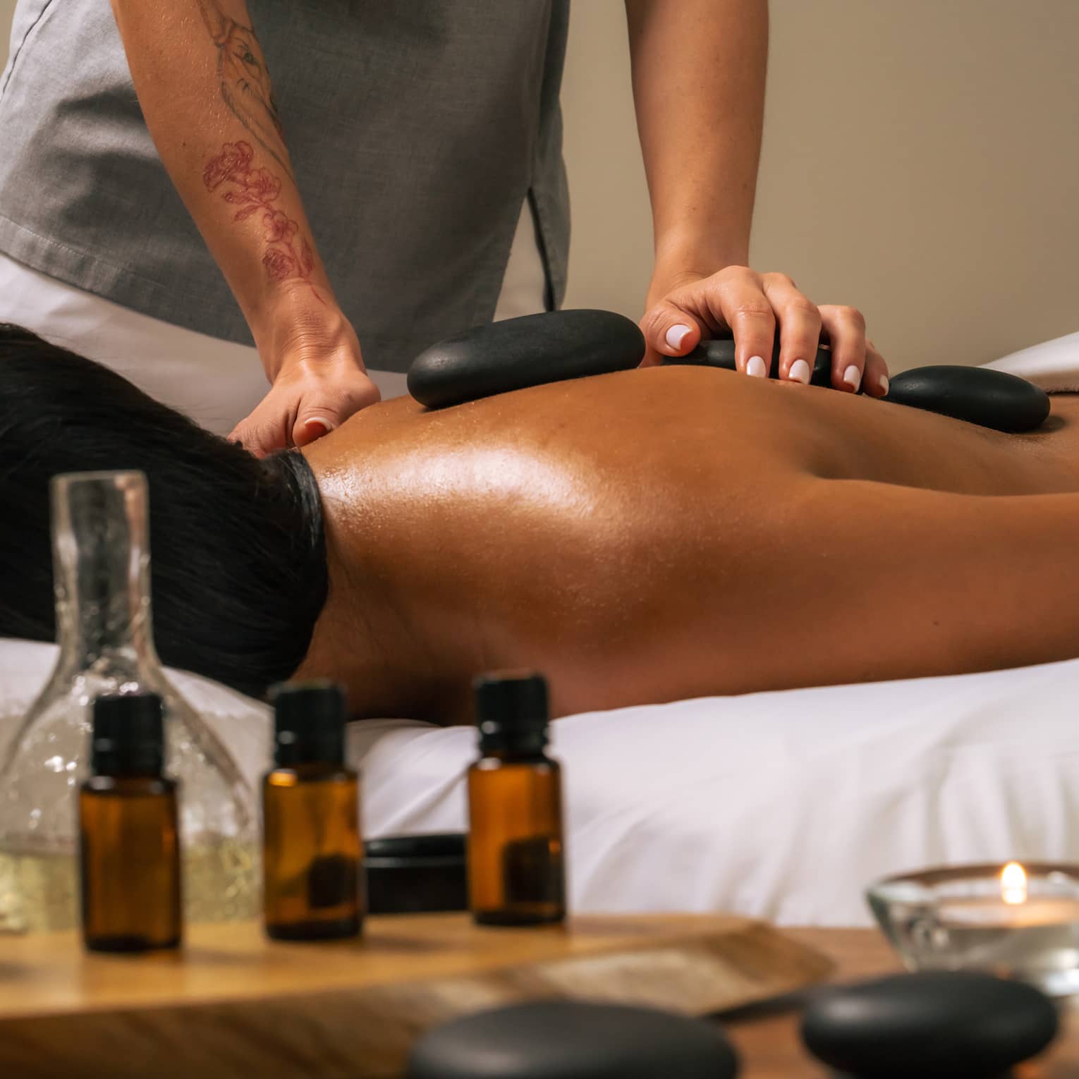 Person lies face down as a massage therapist places black stones on their bare back, with various bottles of oil in the foreground