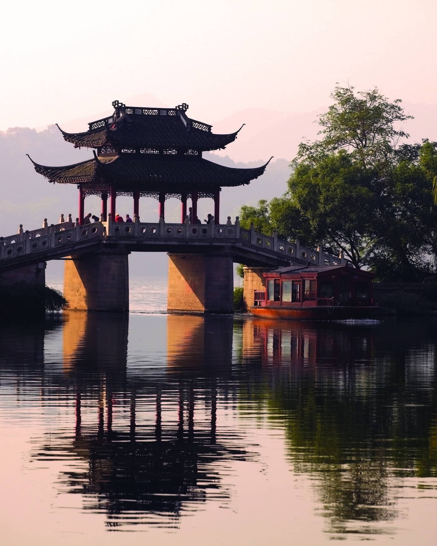 A boat floats towards a stone bridge with a pagoda cover cast in orange light from the setting sun.