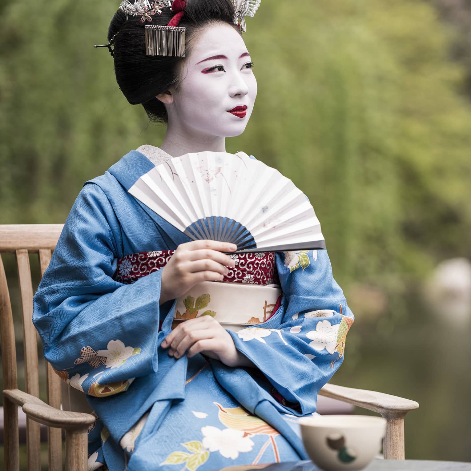 Geiko, a traditional Japanese entertainer, dressed in kimono and holding fan, sitting with cup of tea in garden