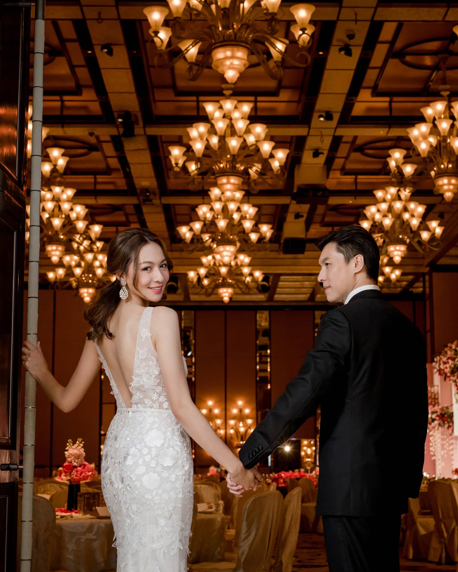 A bride and groom enter through the doors of a grand reception room decorated with banquet tables and ornate chandeliers