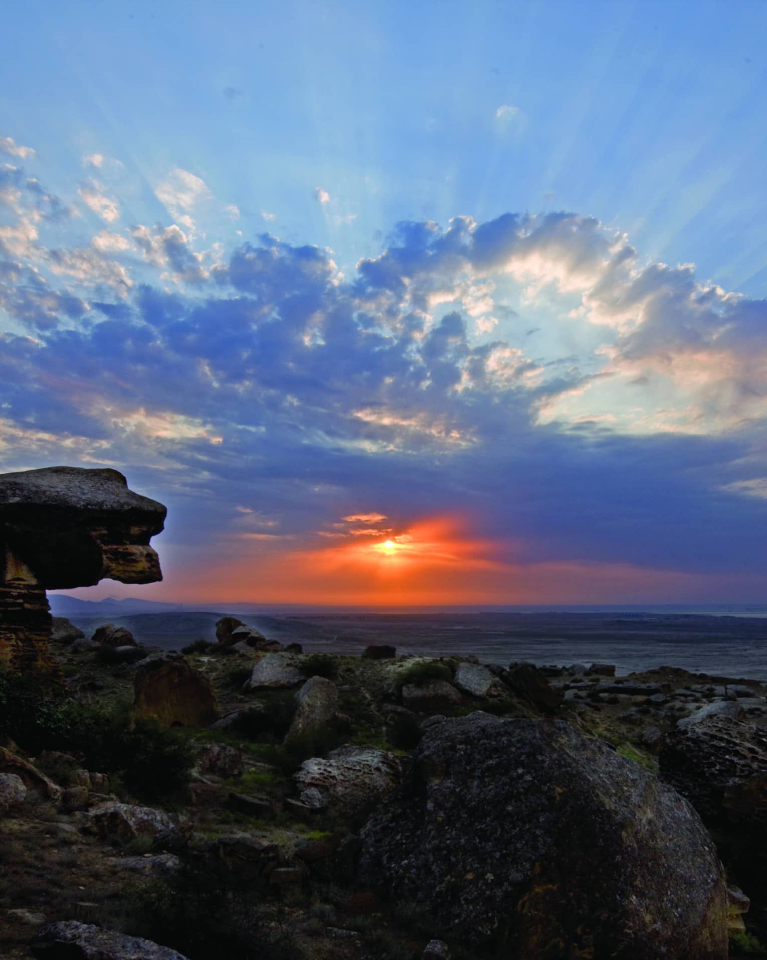 Sunbeams and fluffy azure blue clouds dance across a twilight sky, the vibrant red sun gently illuminating a rocky shoreline.